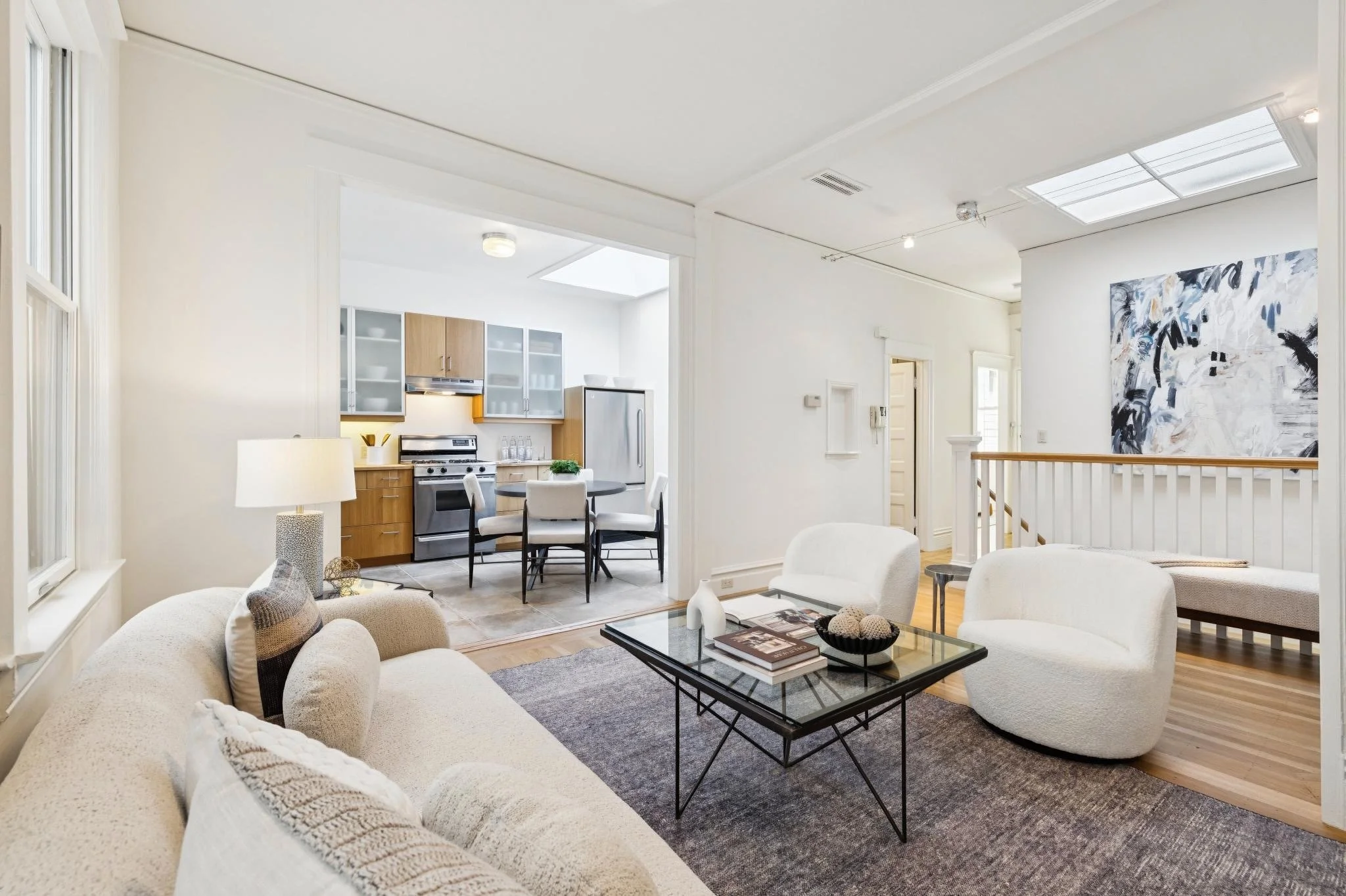 Open-concept living room and kitchen with beige couches, a black coffee table, a dining area, and modern kitchen cabinets with stainless steel appliances, white walls, and hardwood floors.