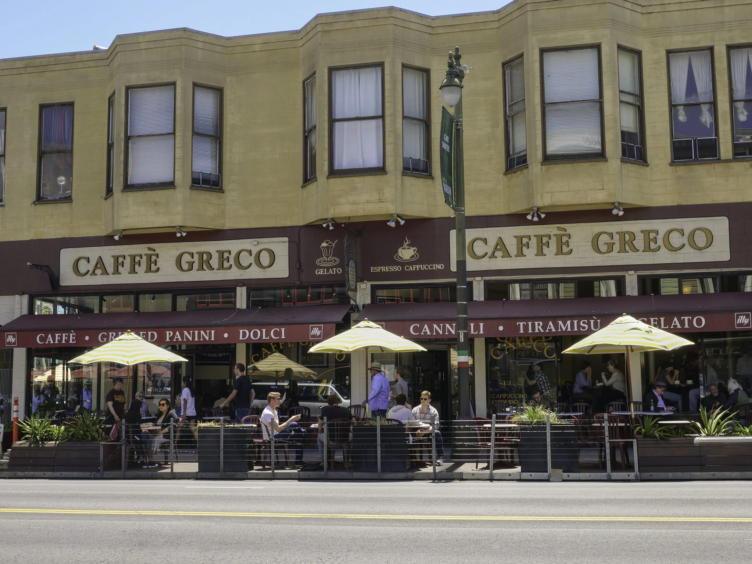 A busy outdoor cafe with people sitting at tables under umbrellas, in front of a yellow building with multiple windows and a sign that reads "Caffé Greco". There is a streetlamp and some greenery in the foreground.
