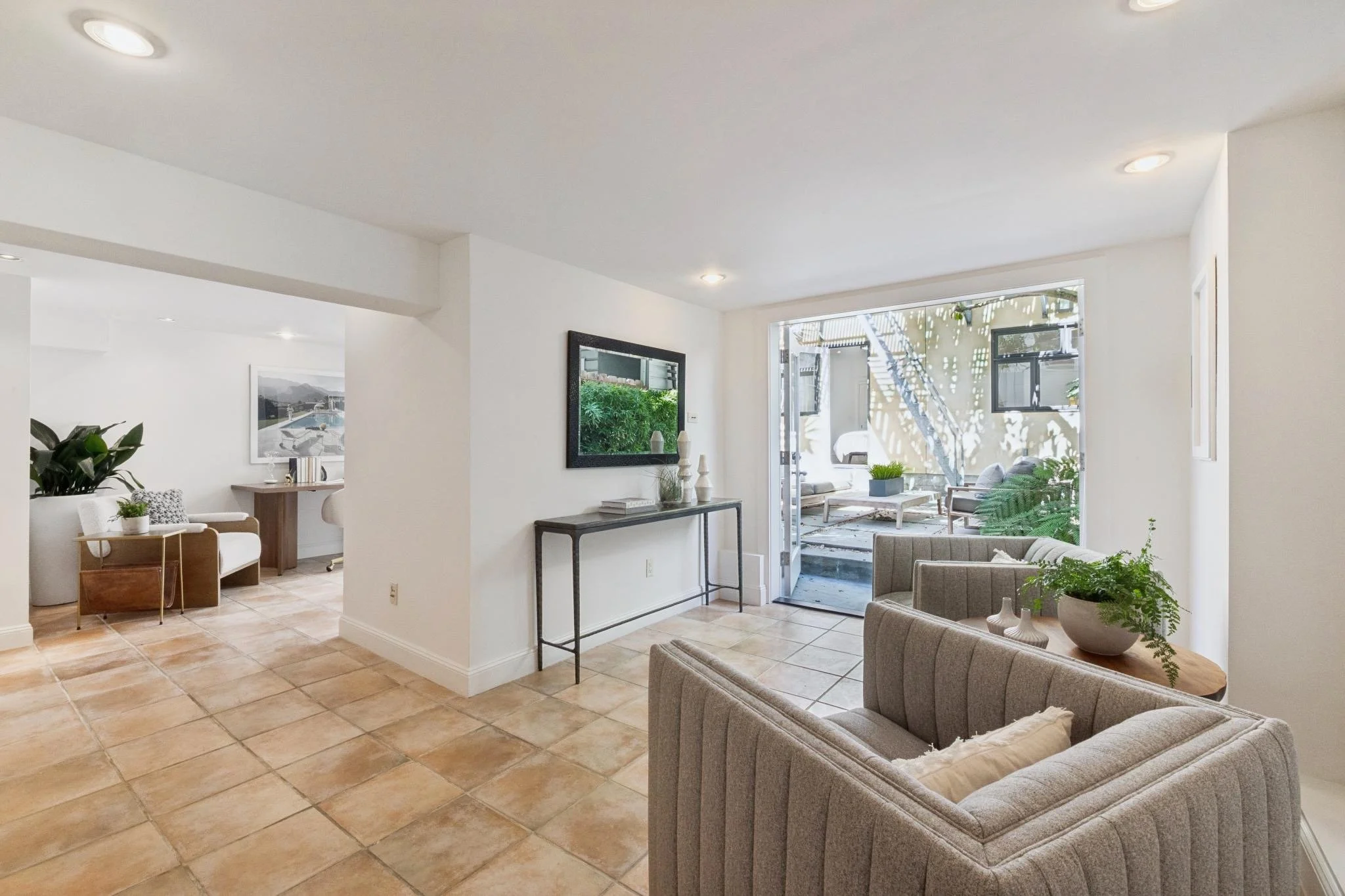 Interior of a living room with beige tiled floor, white walls, and large window leading to an outdoor patio with trees and outdoor furniture.