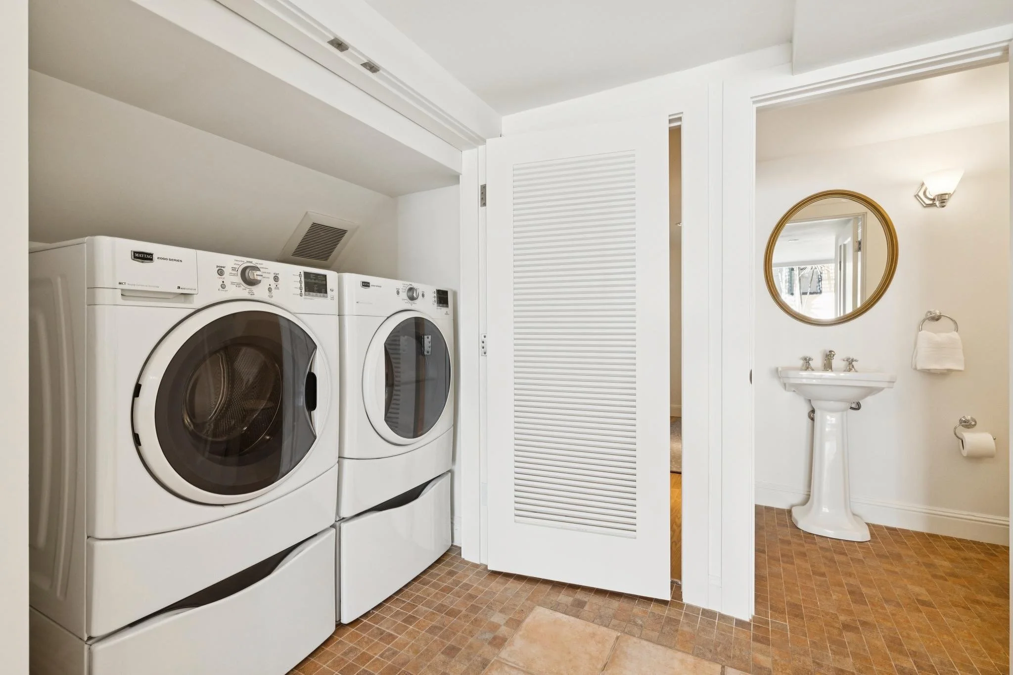 A laundry room with a front load washer and dryer, a louvered closet door, a small bathroom sink with a mirror, and a toilet, all in a neutral color scheme.