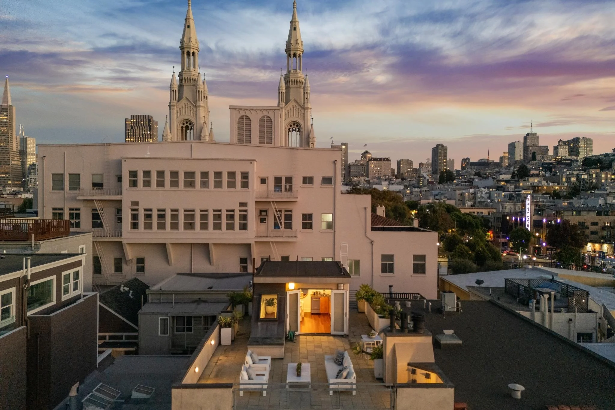 City skyline at sunset with prominent church with twin steeples in foreground, residential rooftops, and city skyscrapers in the background.