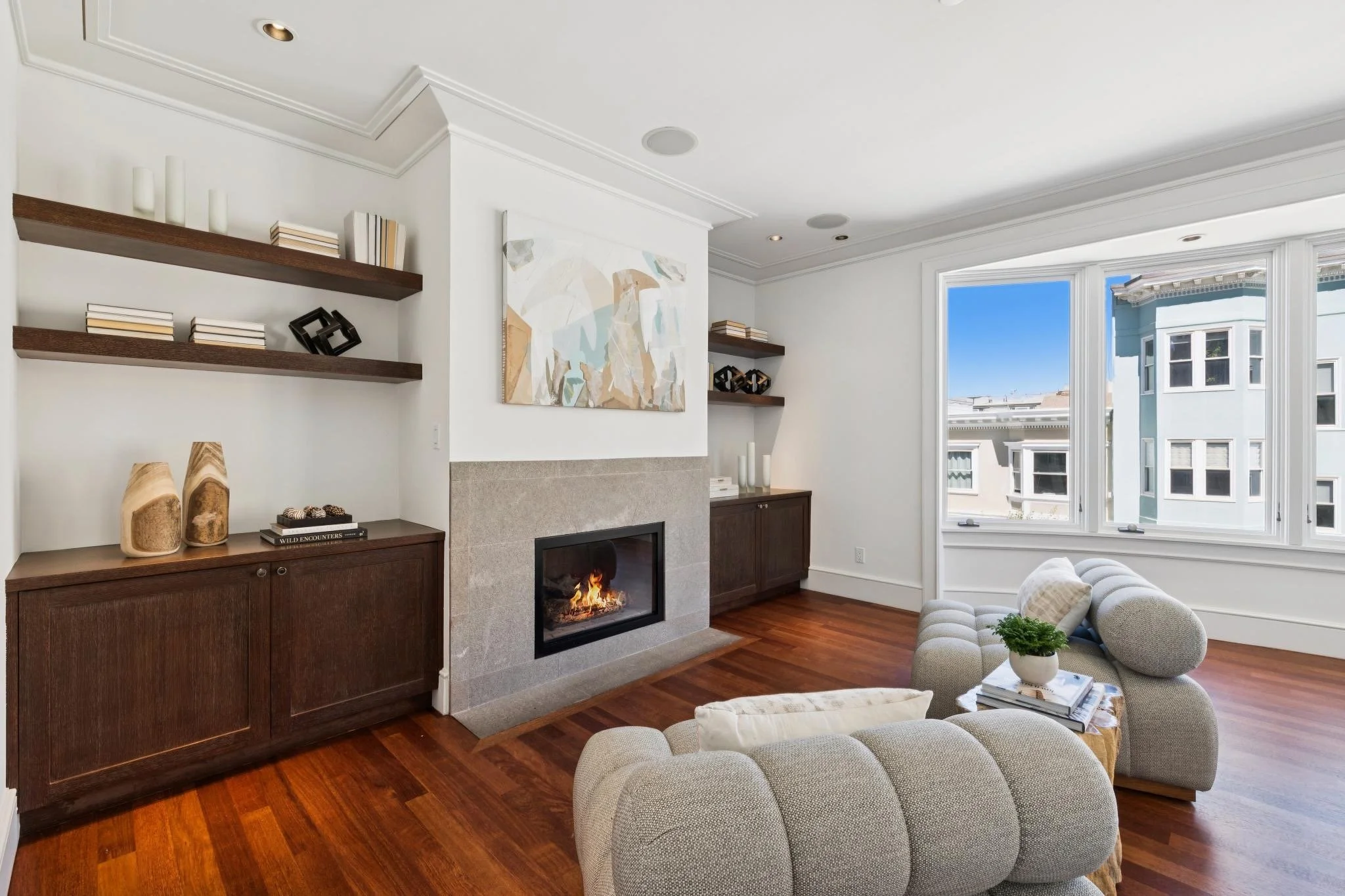 Living room with a fireplace, wooden shelves with books, abstract wall art, large window showing neighboring buildings, and upholstered furniture.