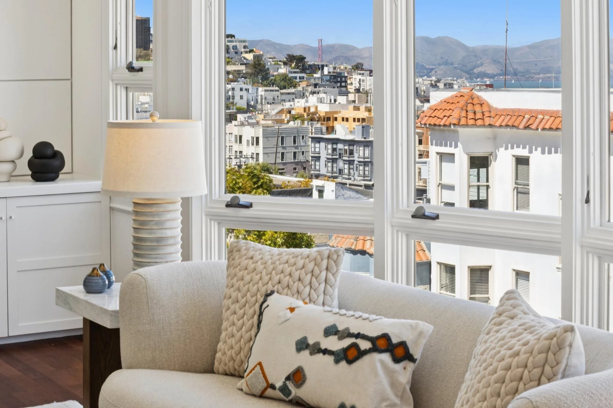 Living room with large windows showing a cityscape with buildings, hills, and a bridge in the distance, white couch with decorative pillows, a side table with vases, and a lamp.