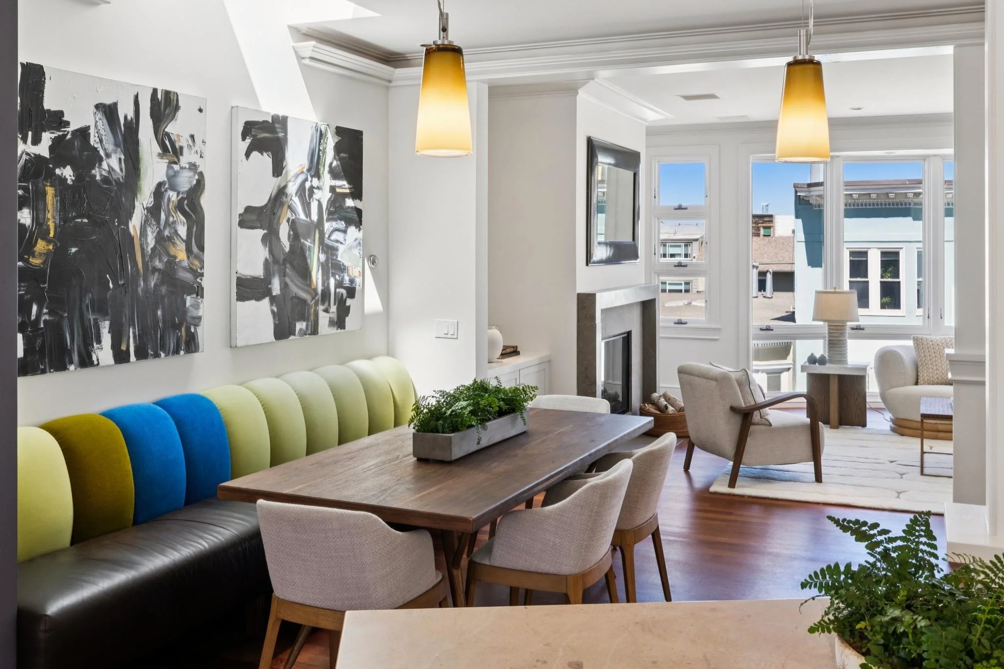 Bright living room with abstract black and white painting on the wall, colorful couch, wooden dining table, and large windows showing neighboring buildings and a blue sky.