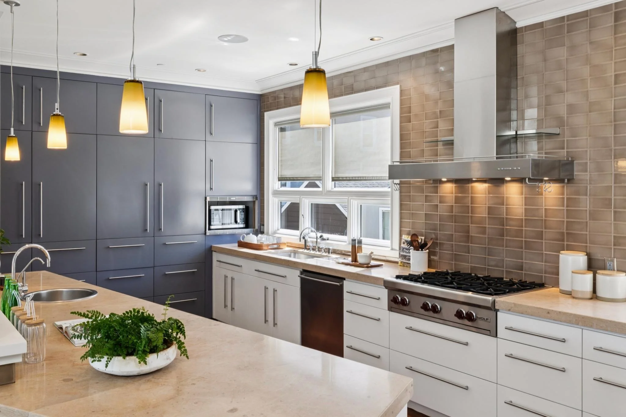 Modern kitchen with gray cabinets, white countertops, and beige tile backsplash. Pendant lights hang over the island, which features a potted green plant and a book. There are three windows with blinds above the sink, and kitchen appliances such as a