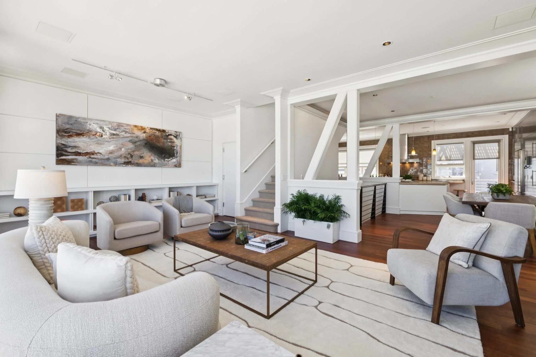 Living room with white sofa, armchairs, coffee table, wall art, and plants, in an open-plan space with a staircase and kitchen in the background.