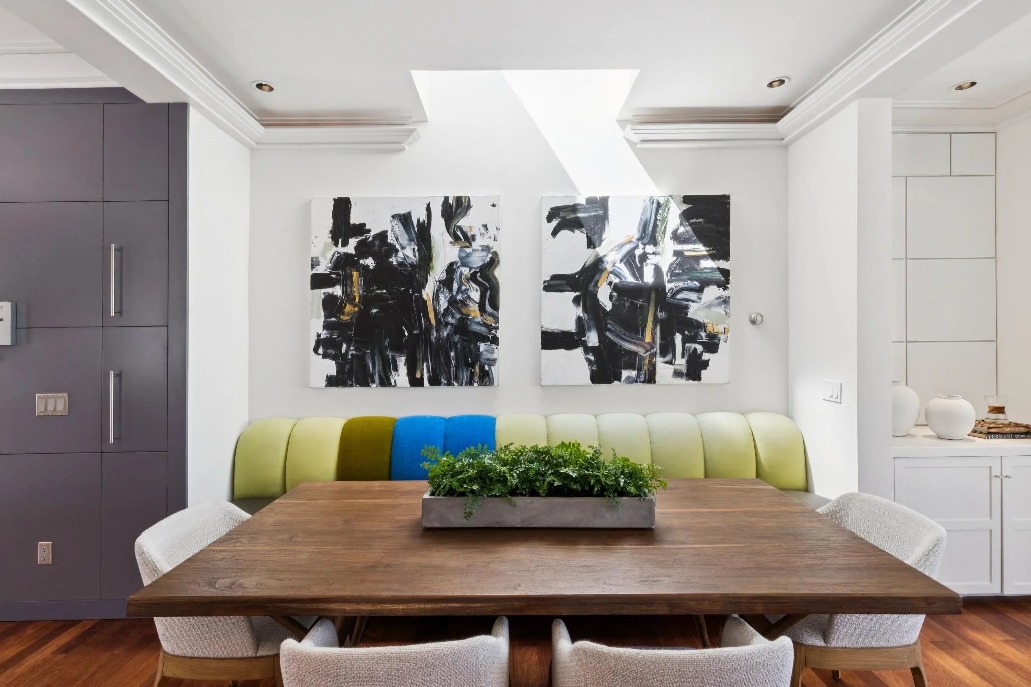 A dining room with a wooden table, six chairs, a green and blue sofa, a hanging art piece with abstract black and white design, a plant centerpiece, and built-in storage cabinets.