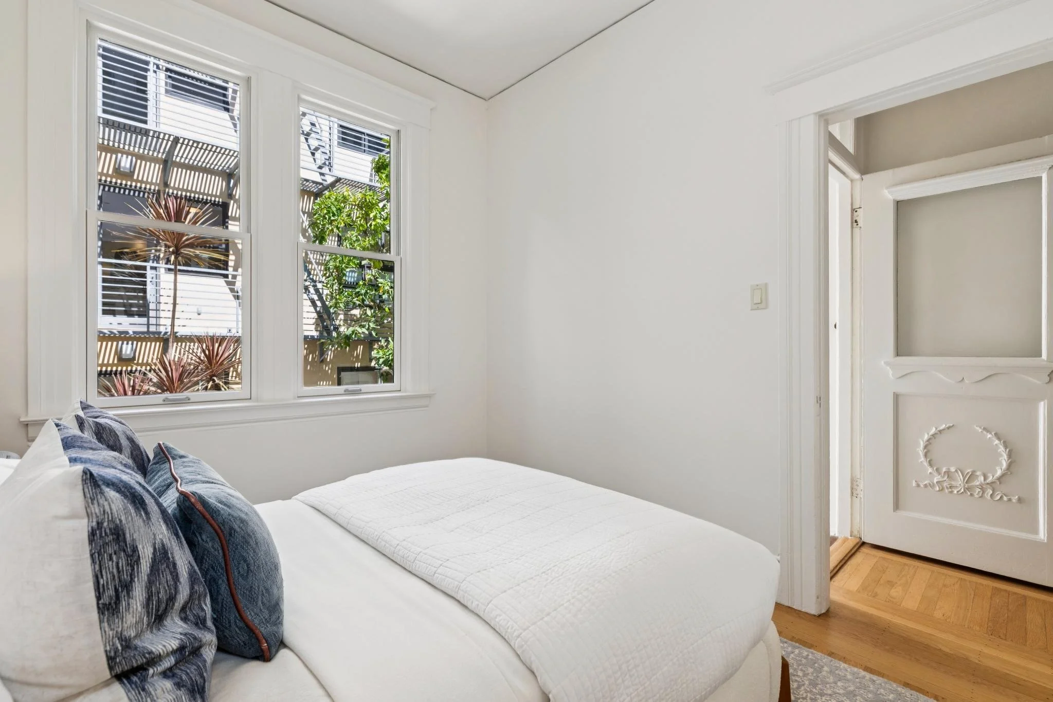 A bedroom with two large windows showing outdoors plants and trees. The bed has white covers and dark blue patterned pillows. The room has white walls, a hardwood floor, and an ornate white door.