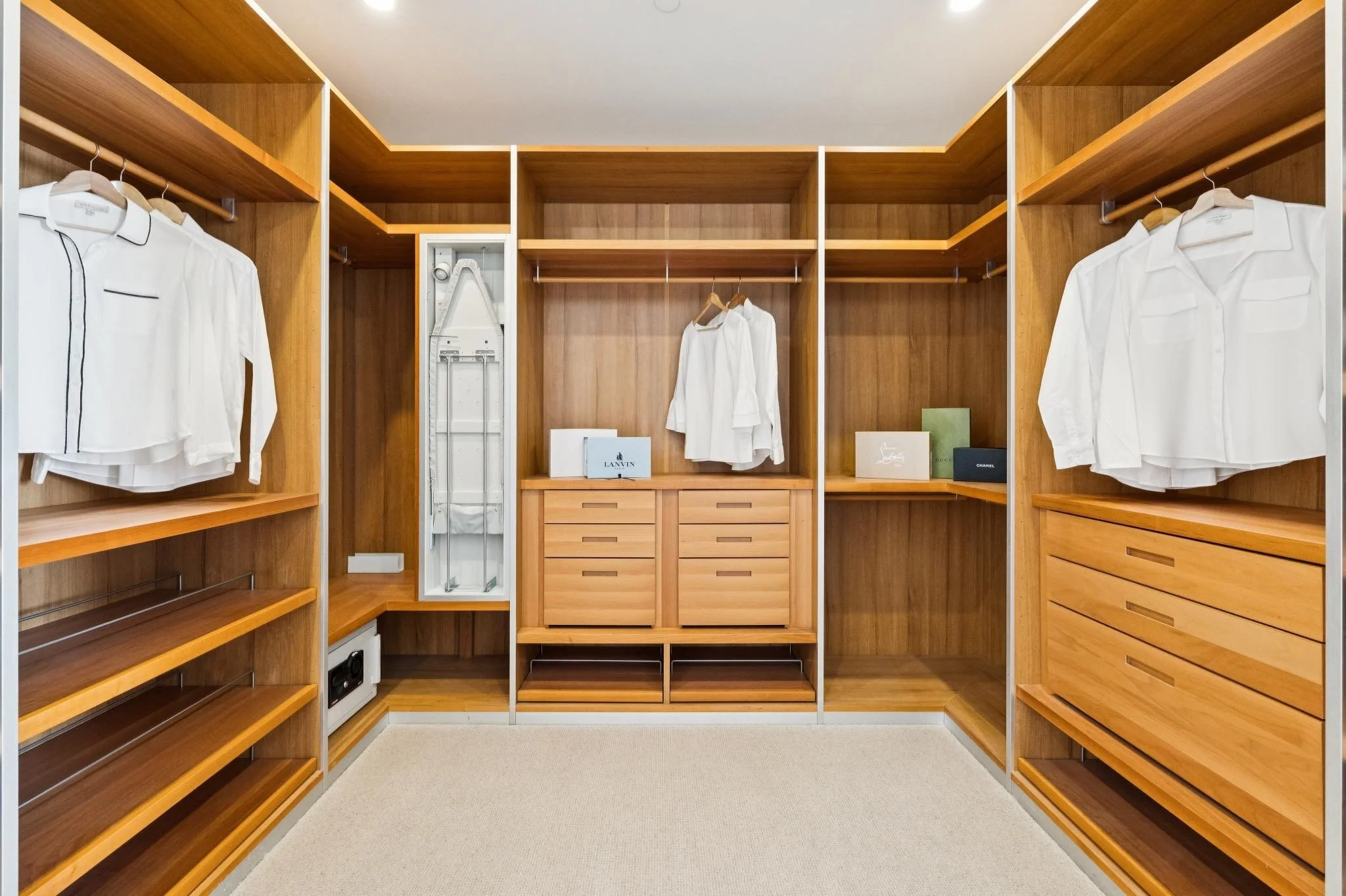 A walk-in closet with wooden shelving and drawers, white shirts hanging on hangers, a suitcase, and several designer bags on display.