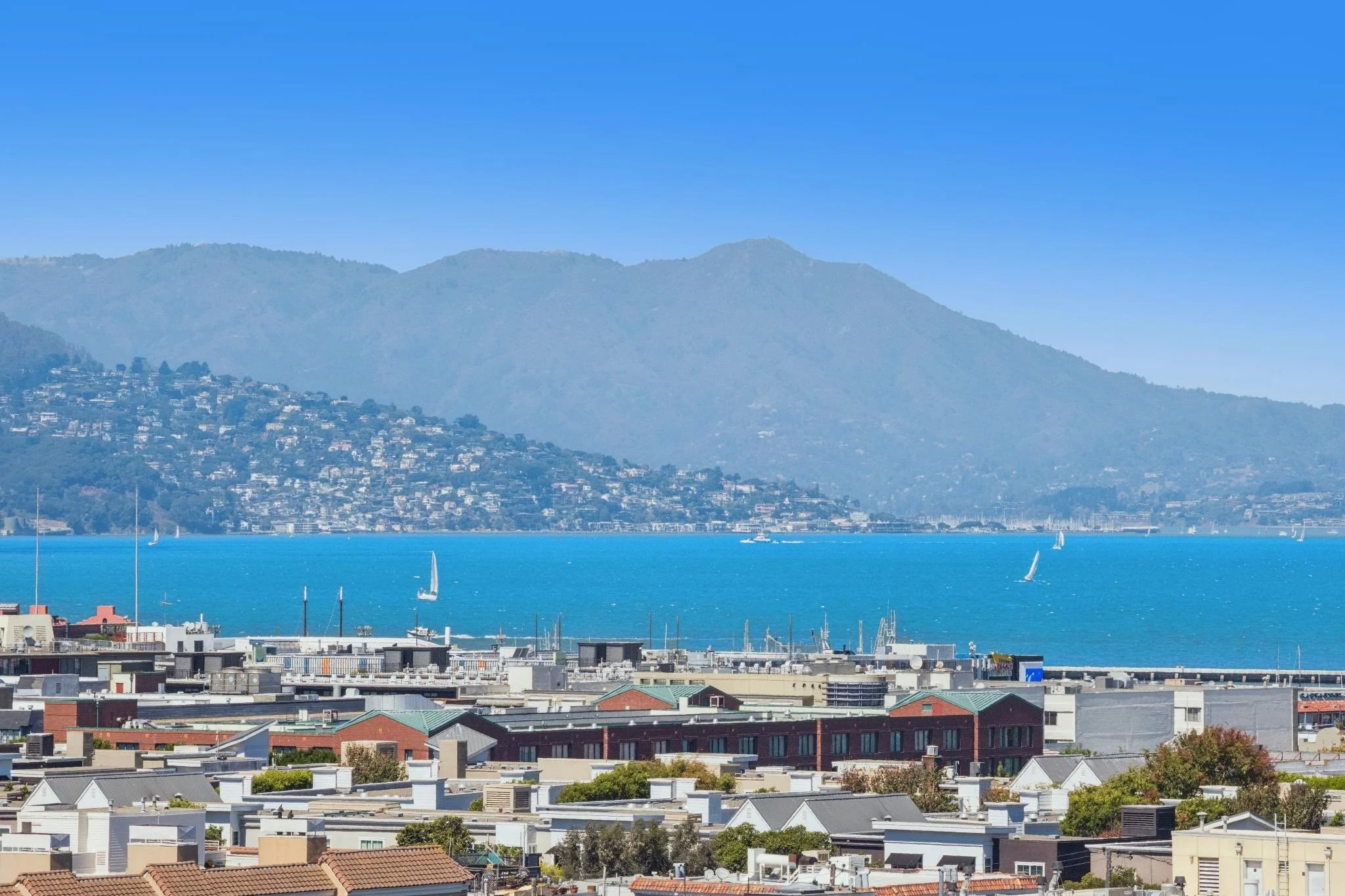 Cityscape with residential buildings in the foreground, water with sailboats in the middle ground, and a mountain range in the background under a clear blue sky.