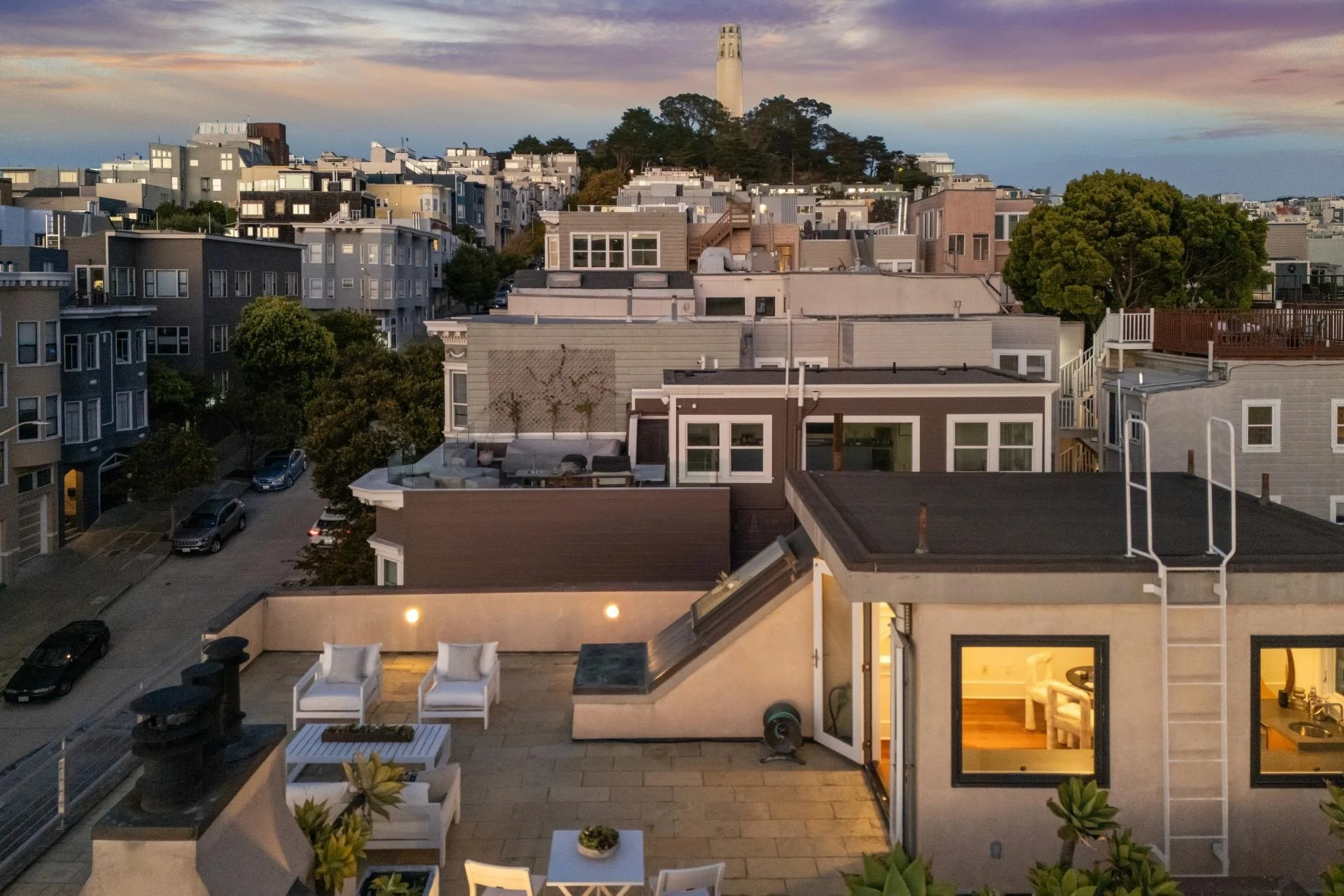 View of a residential neighborhood during sunset with many apartment buildings and houses, some with decks and outdoor furniture, and a tall monument or tower on a hill in the background, with trees and a colorful sky.
