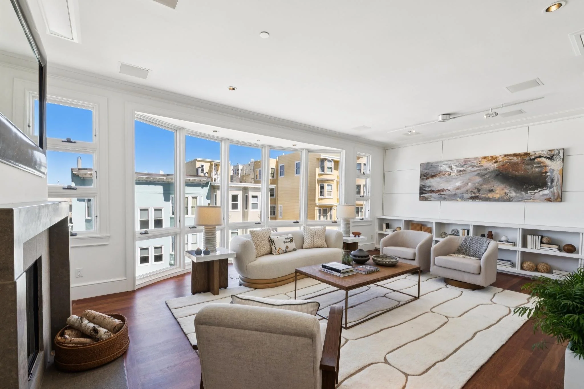Bright living room with large bay windows, white walls, modern artwork, beige sofa, armchair, coffee table, shelves, and decorative items.