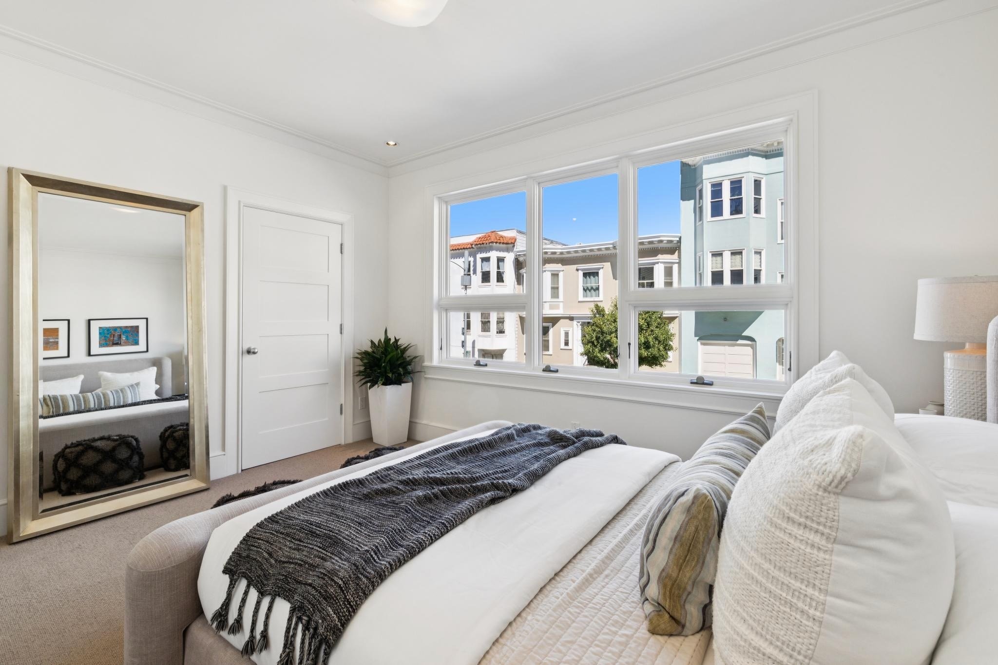 A cozy bedroom with a large window showing neighboring houses and blue sky. The bed has white and striped pillows, and a black and white striped throw. There is a gold-framed mirror and a potted plant near the window.