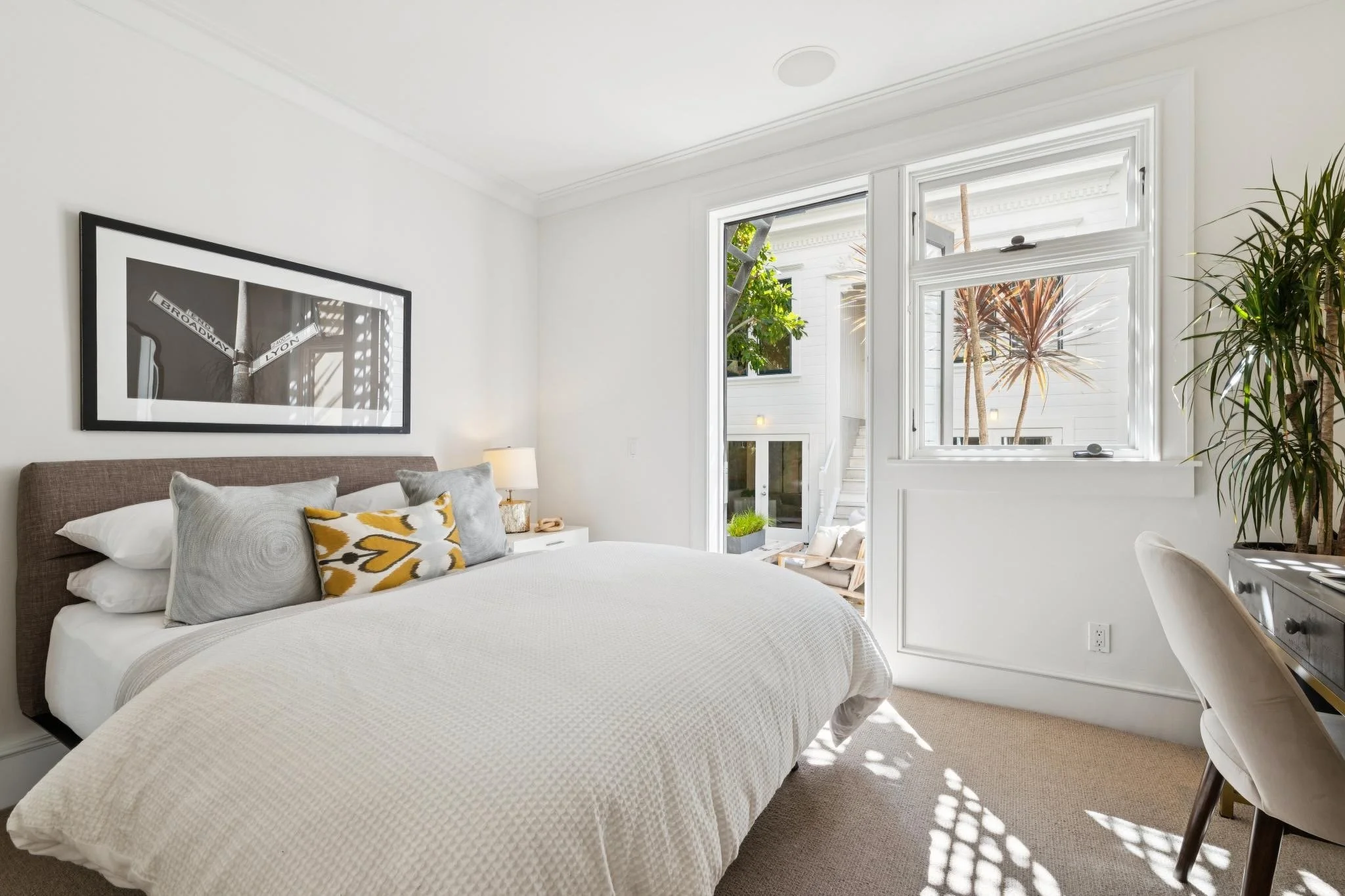 Bright bedroom with a bed featuring white and gray pillows, large windows with sunlight streaming in, a potted plant, and a black and white framed artwork on the wall.