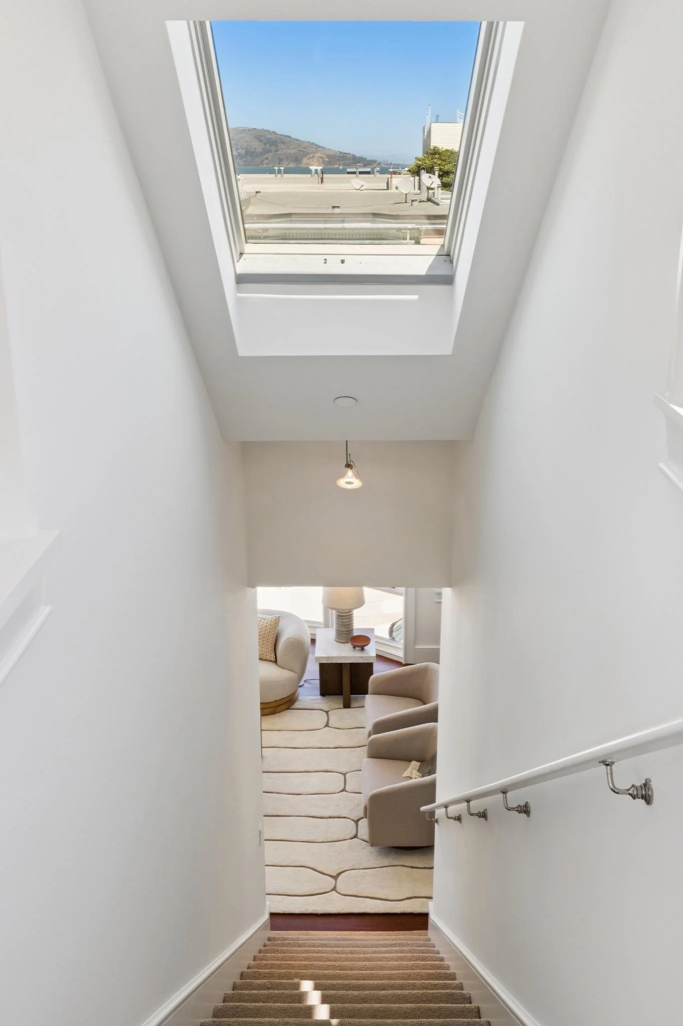 Looking down a staircase at the bottom of a hallway with a view of a living room with beige chairs, a rug, and a white wall. A skylight at the top shows a clear sky and a mountain in the distance.