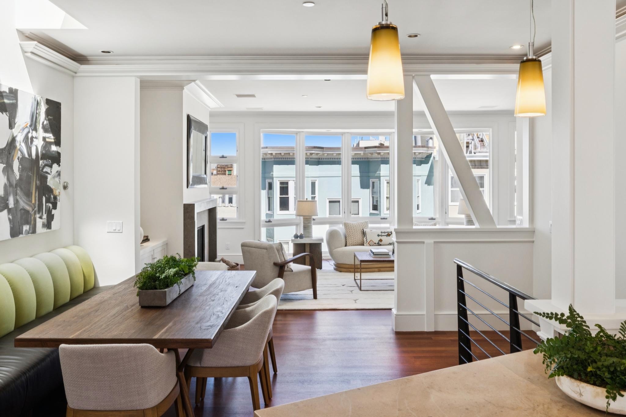 A spacious, modern living room with large windows, white walls, and contemporary furniture, including a dining table with chairs, a sitting area with armchairs, and hanging pendant lights.