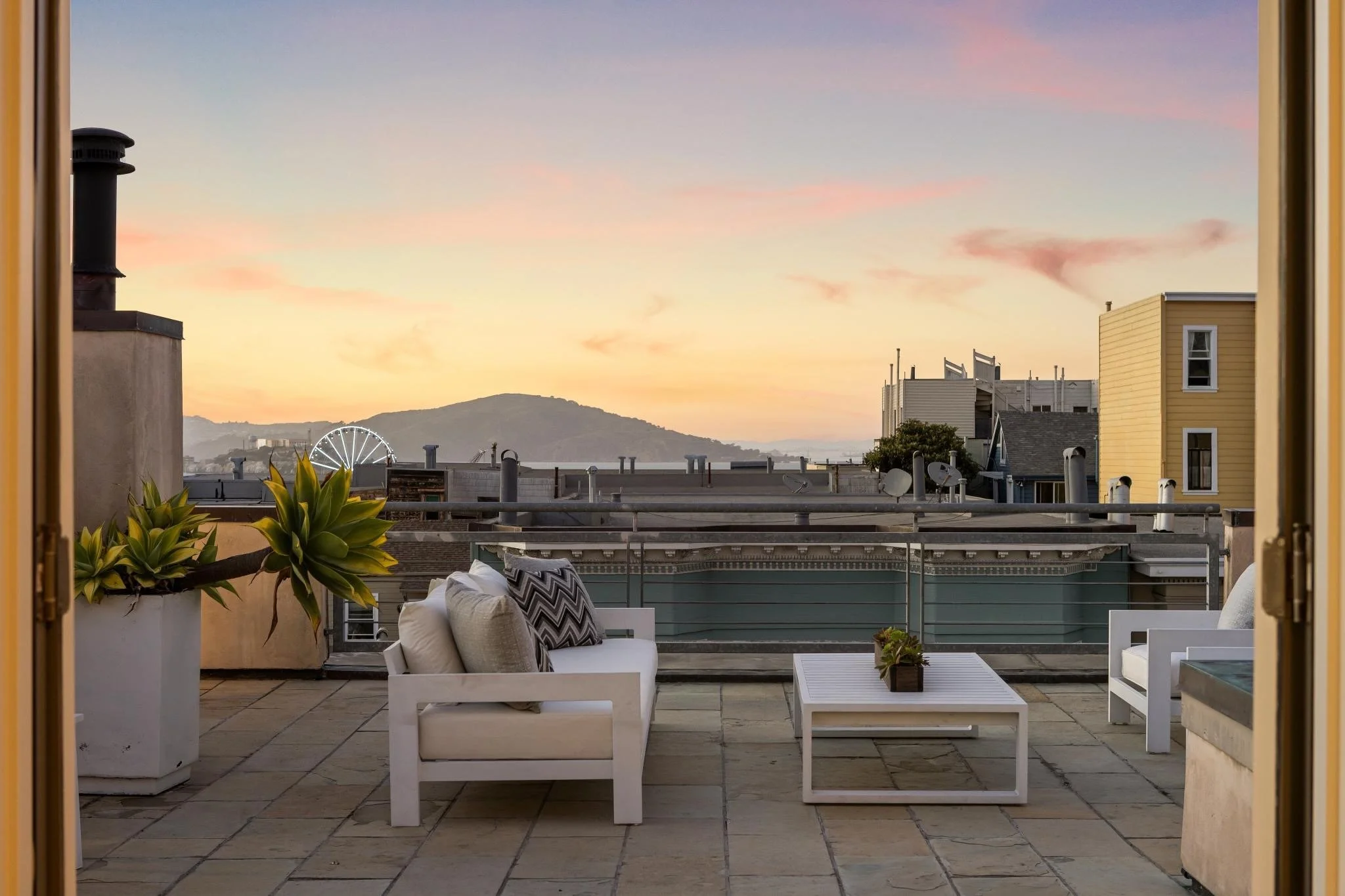 A rooftop patio at sunset with outdoor seating, potted plants, and a view of distant hills and a Ferris wheel.