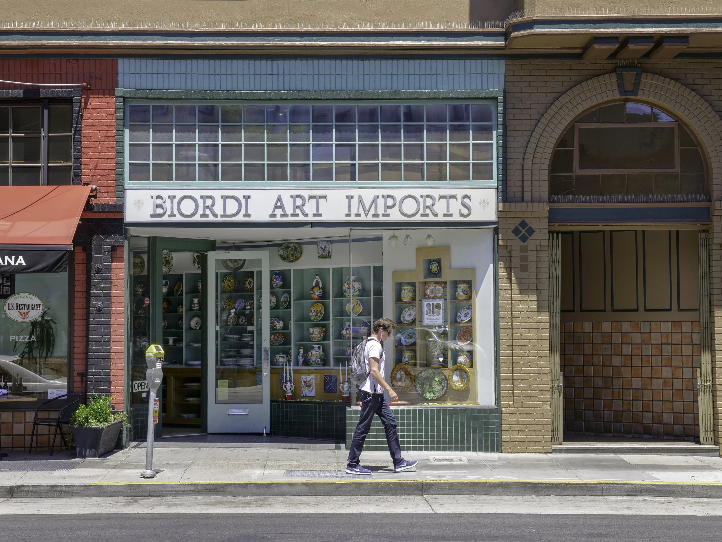 Colorful storefront with large window display of decorative plates and artwork, sign reading "Biordi Art Imports," and a person walking on the sidewalk in front.
