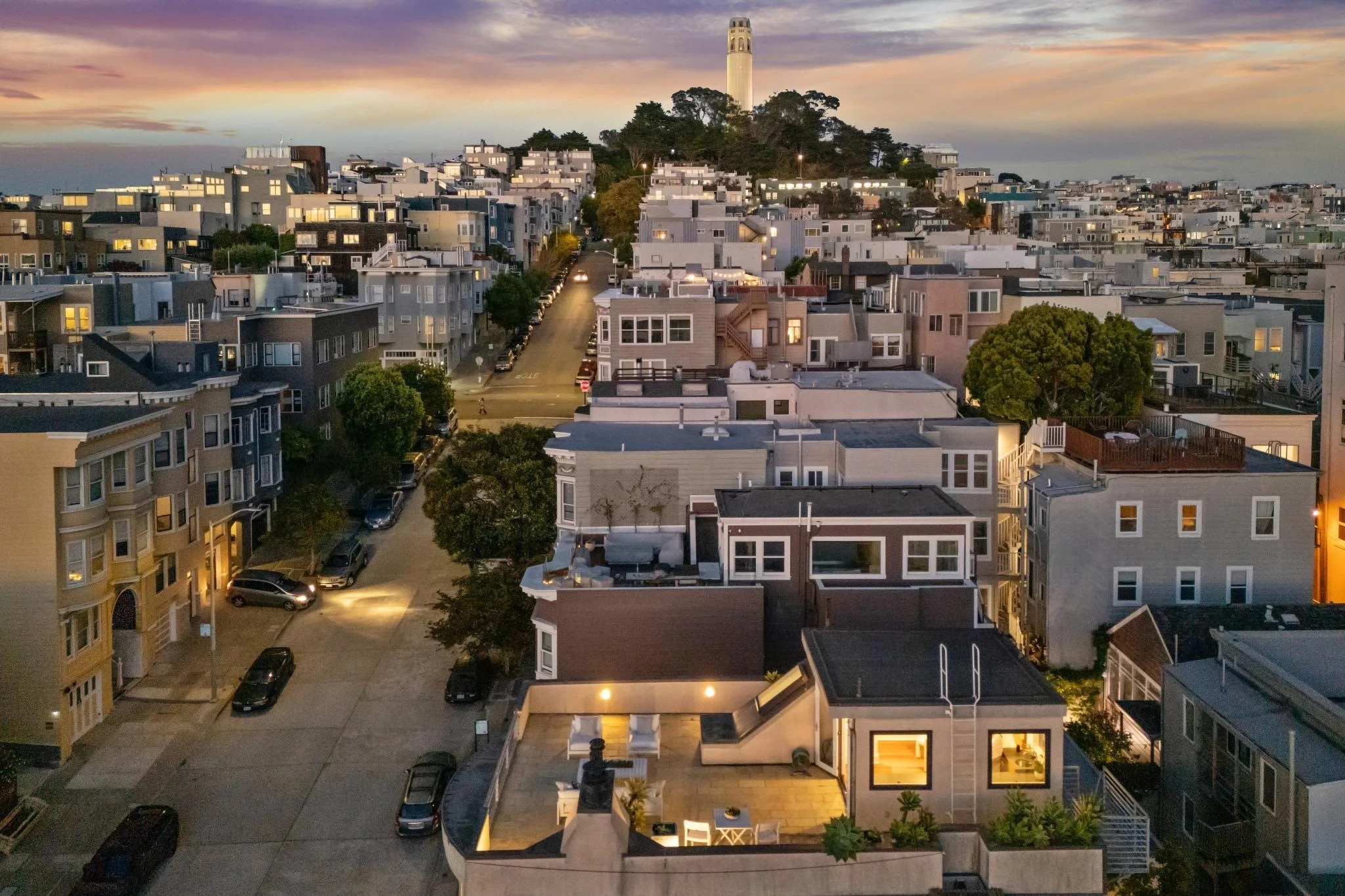 An aerial view of a San Francisco neighborhood at dusk, showing closely packed houses, streets with parked cars, a hill with trees, and a prominent tower in the distance under a sunset sky.