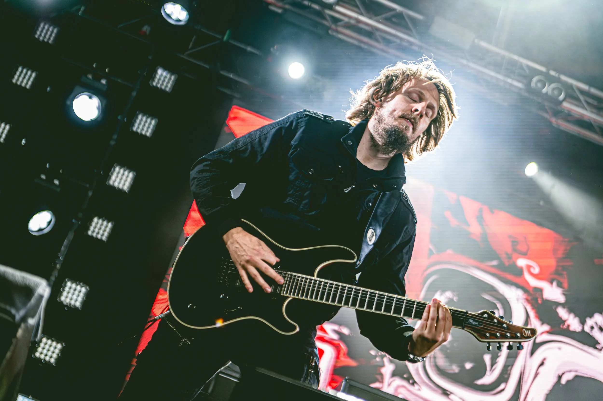ONLY HUMAN guitarist Martin Hastrup, with long hair and a beard, playing an electric guitar on stage, with bright stage lights and a dark background.