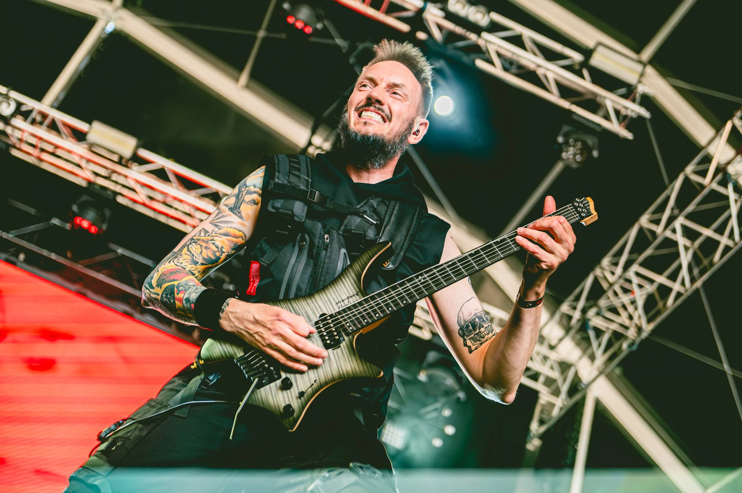 ONLY HUMAN guitarist Jack Kijne, with tattoos and a beard playing an electric guitar on stage during a concert, smiling, with stage equipment and lighting structures in the background.