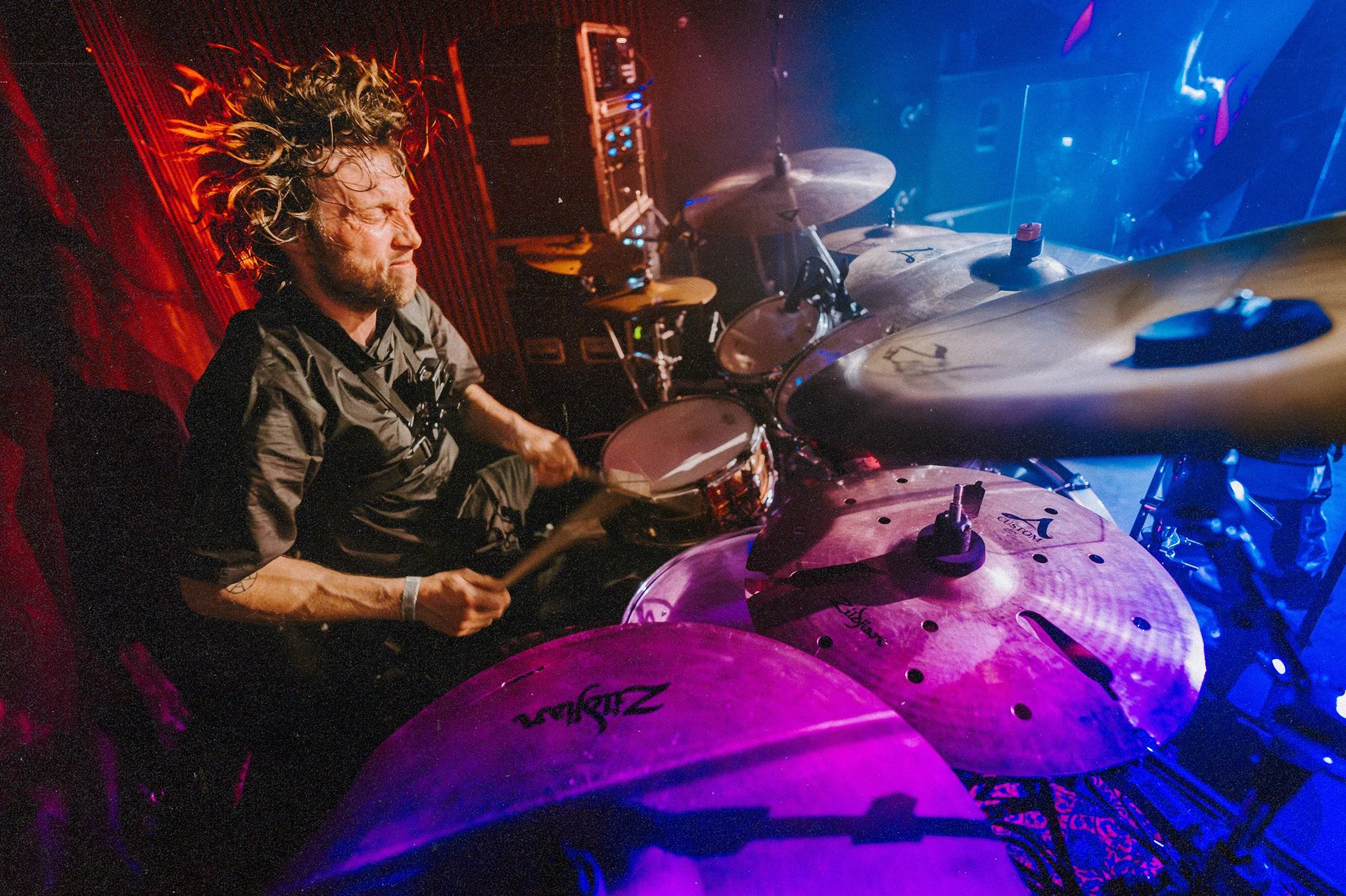 ONLY HUMAN drummer Andreas Pröll with long, curly hair is performing on stage, surrounded by red and blue stage lighting, with a drum set and electronic equipment in the background.