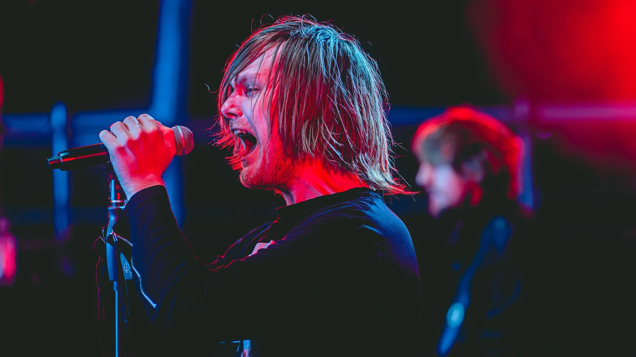 ONLY HUMAN frontman Patrick Christensen, passionately singing into a microphone on stage, illuminated by red and blue stage lights, with member Martin Hastrup blurred in the background.
