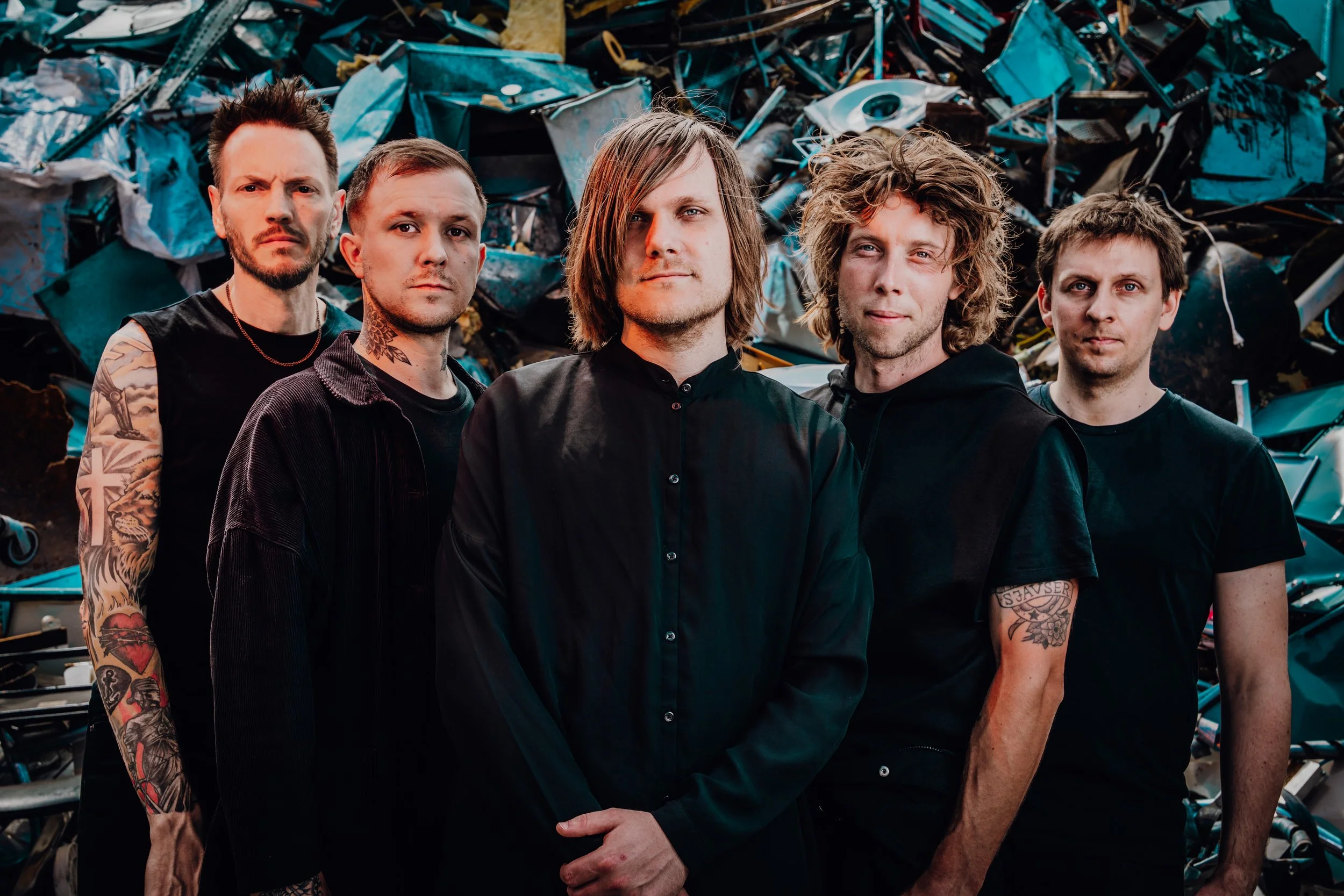 Press photo of the five members of ONLY HUMAN with tattoos and casual clothing standing in front of a pile of scrap metal. Left-to-right: Jack, Guillaume, Patrick, Andreas, Martin.
