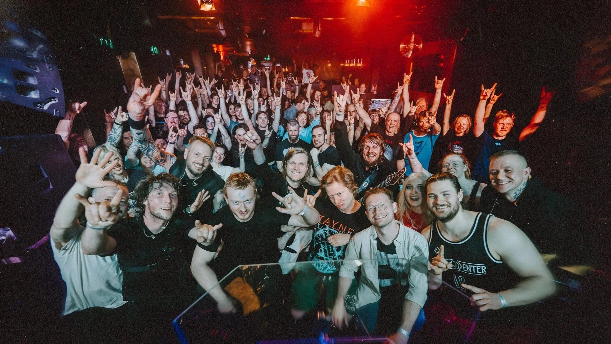 Photo taken from the stage overlooking a group of people at a concert or event smiling, raising hands, and making rock and roll gestures in a live venue setting.
