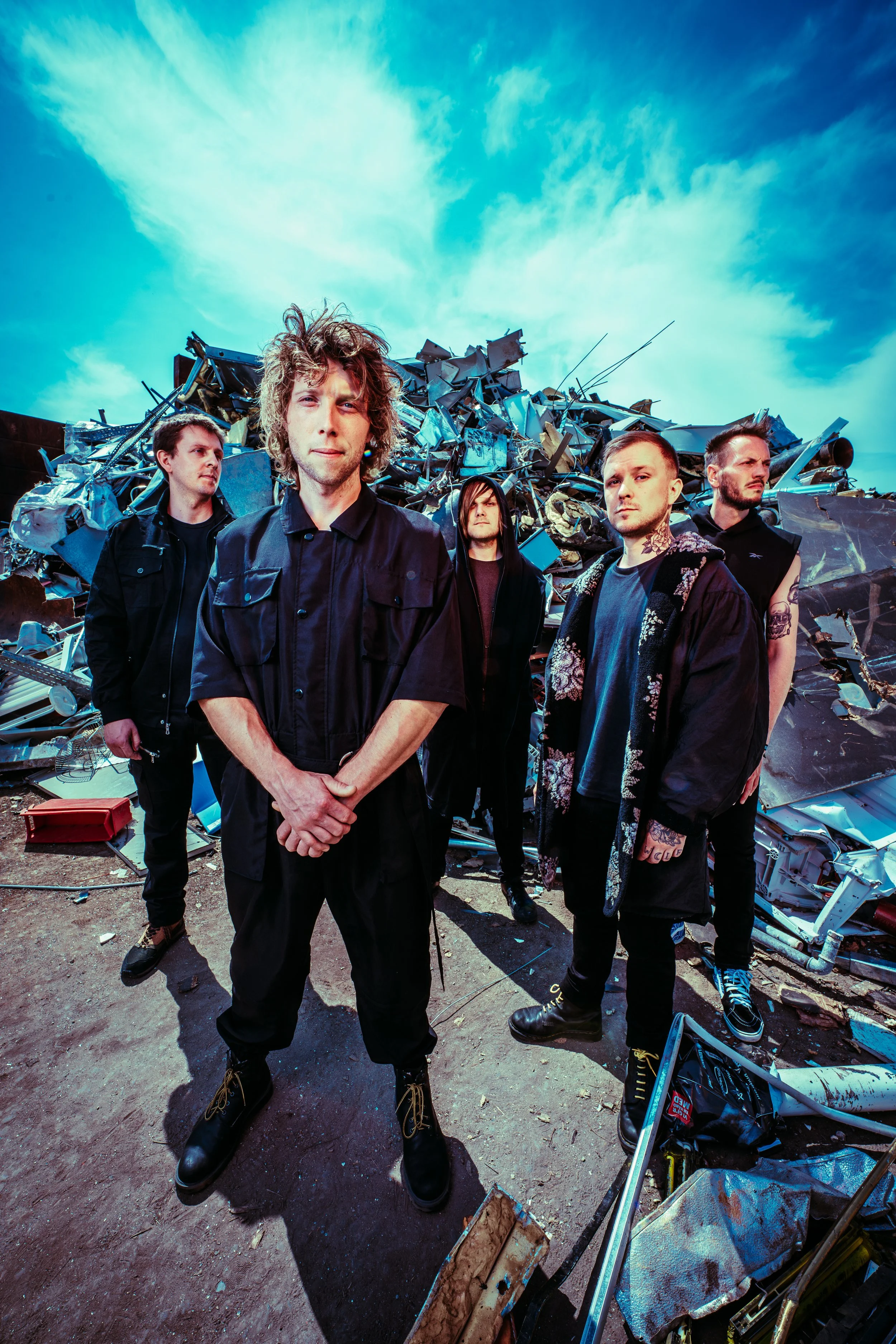Press photo of the five members of ONLY HUMAN standing in front of a large pile of scrap metal and debris on a dirt ground, under a blue sky with some clouds. Left-to-right: Martin, Andreas, Patrick, Guillaume, Jack.