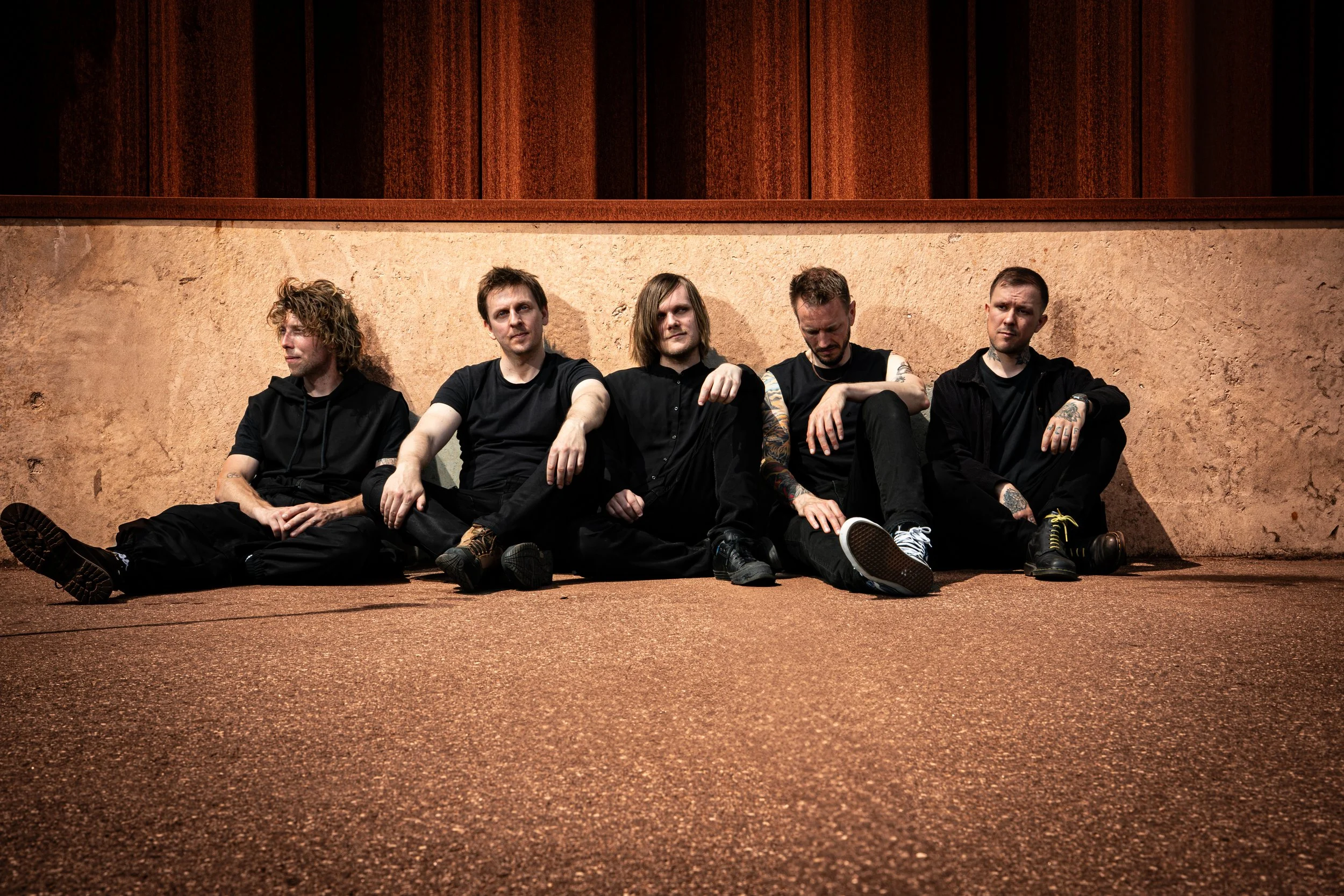 The five members of ONLY HUMAN sitting on the ground against a beige wall with dark wooden paneling above, dressed in black casual clothing, some with tattoos. Left-to-right: Andreas, Martin, Patrick, Jack, Guillaume.
