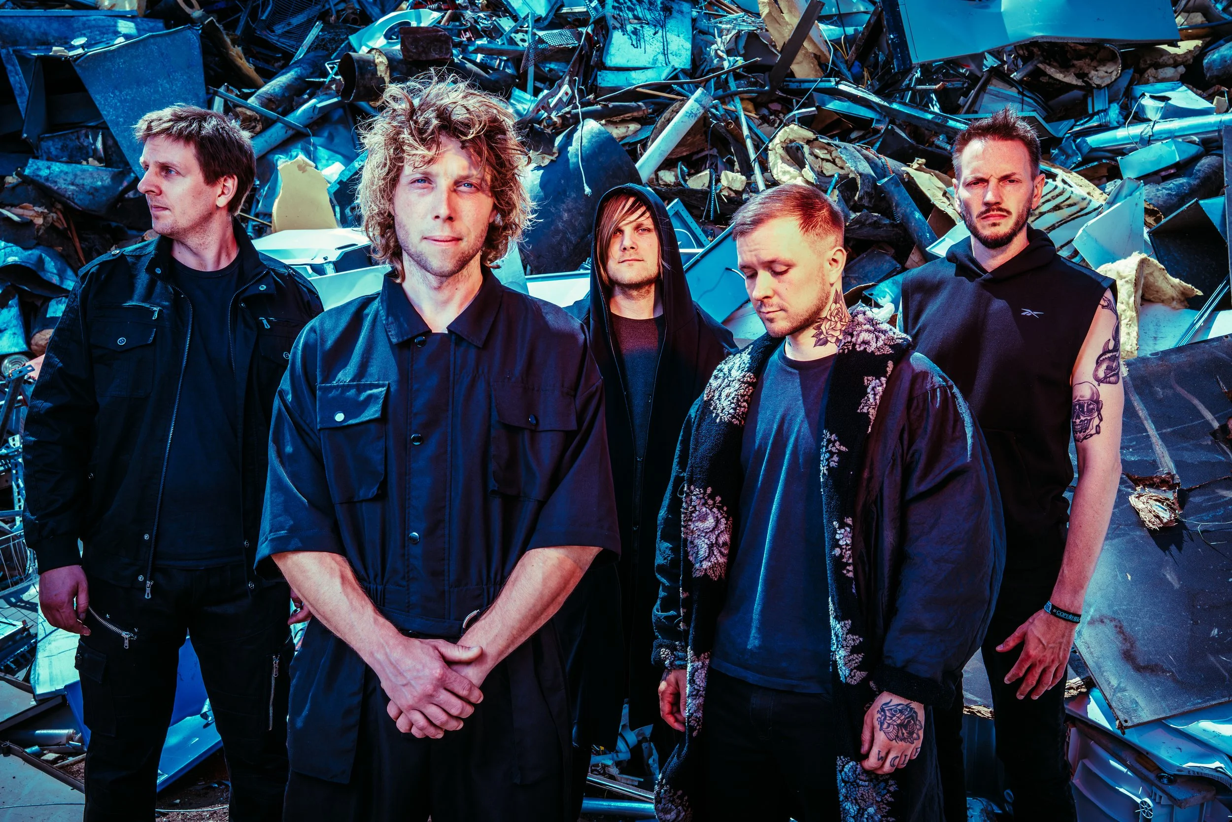 Press photo of the five members of ONLY HUMAN standing in front of a large pile of scrap metal and debris. Left-to-right: Martin, Andreas, Patrick, Guillaume, Jack.