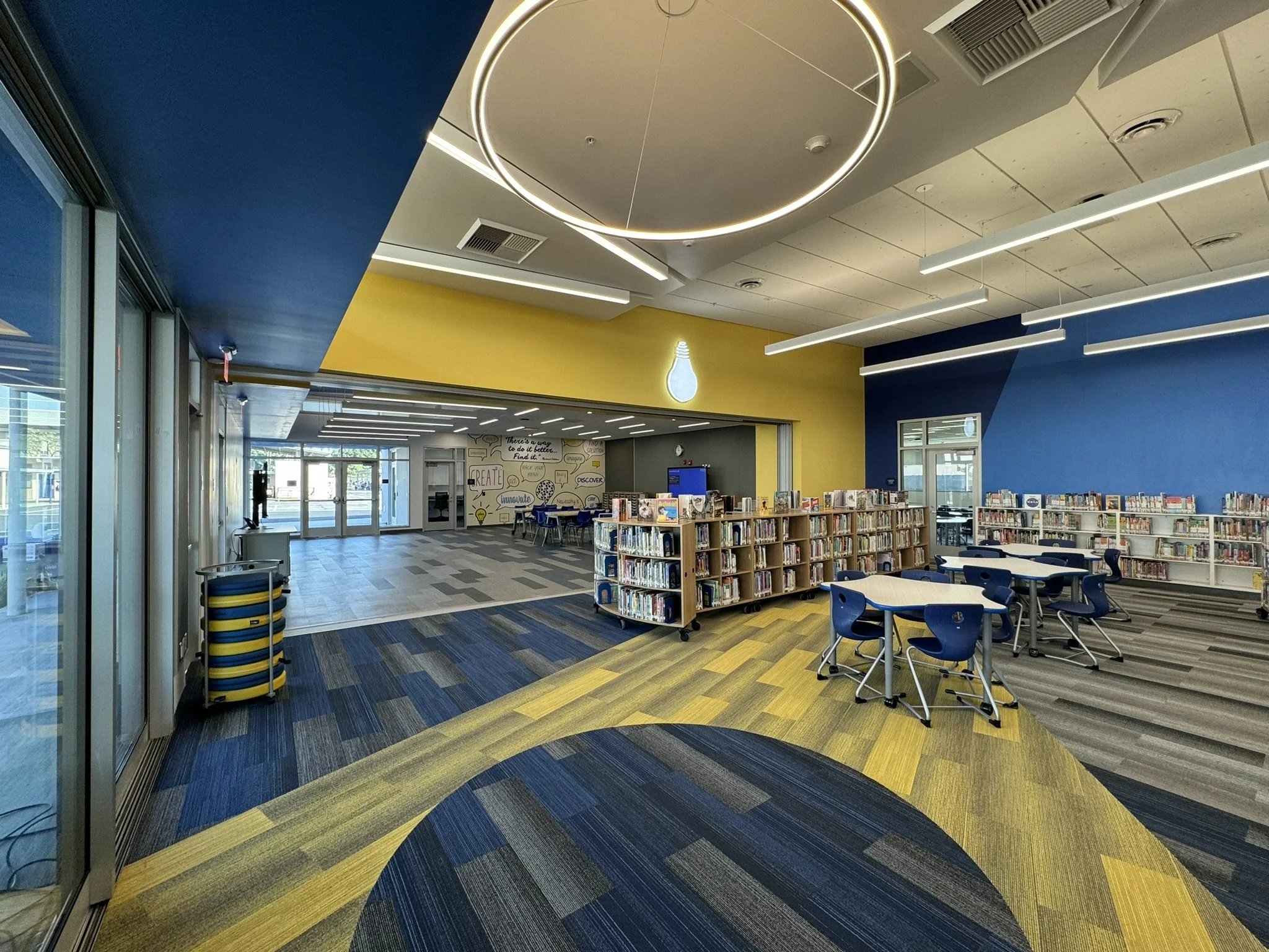 Interior of a modern library with colorful patterned carpet, bookshelves, and blue chairs, with a yellow accent wall featuring a light bulb graphic and quote.