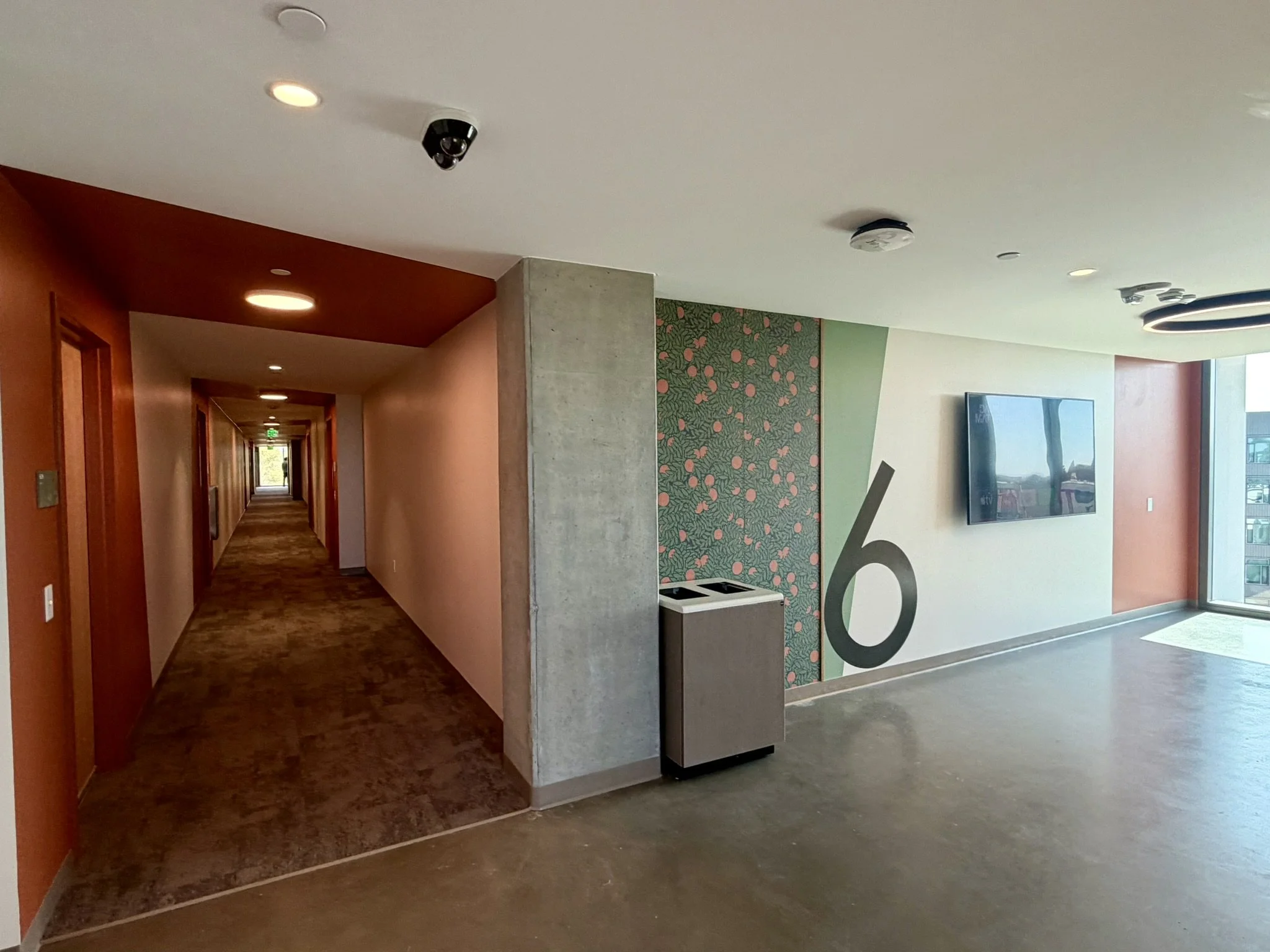 Interior of a modern building lobby or corridor with a wall-mounted television, decorative wallpaper, and ceiling lights, with a hallway extending into the distance.