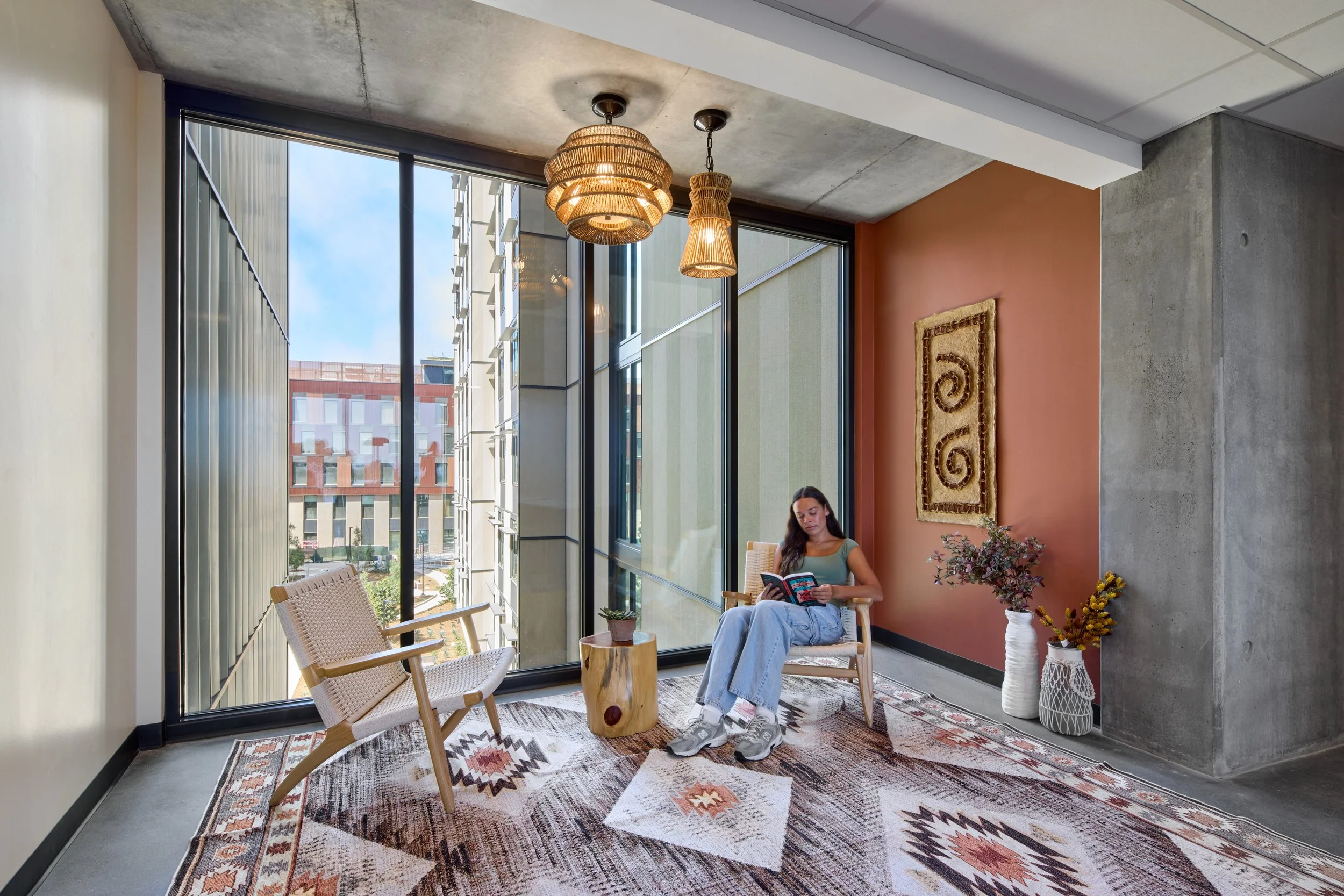 A woman sitting in a modern indoor space, reading a book, with large window views of a cityscape in the background. The decor includes woven pendant lights, a patterned rug, potted plants, and contemporary furniture.