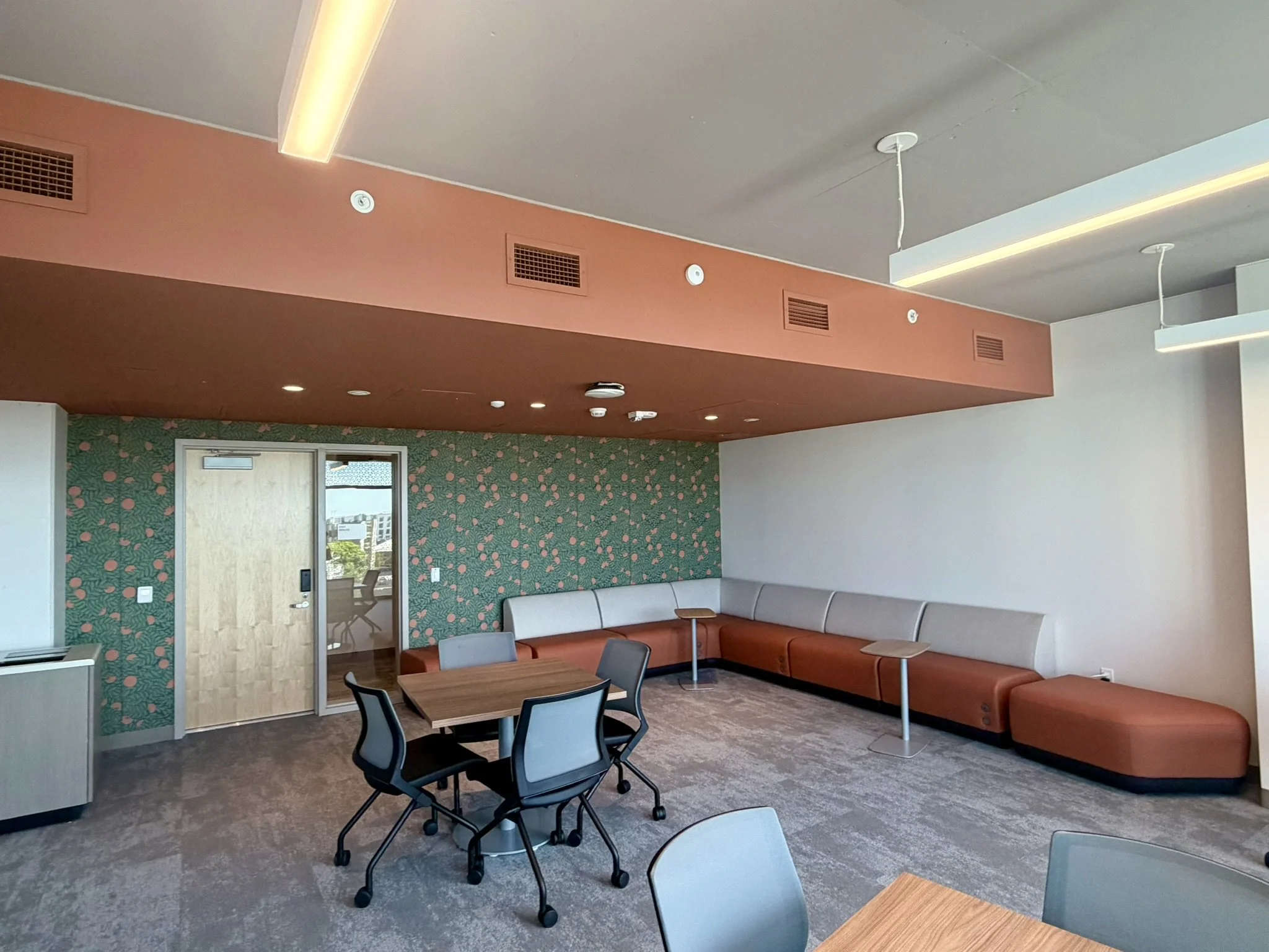 Commercial lounge area with wood and black chairs around tables, a patterned wallpapered wall, a tan corner sofa, and a ceiling with linear lighting fixtures.