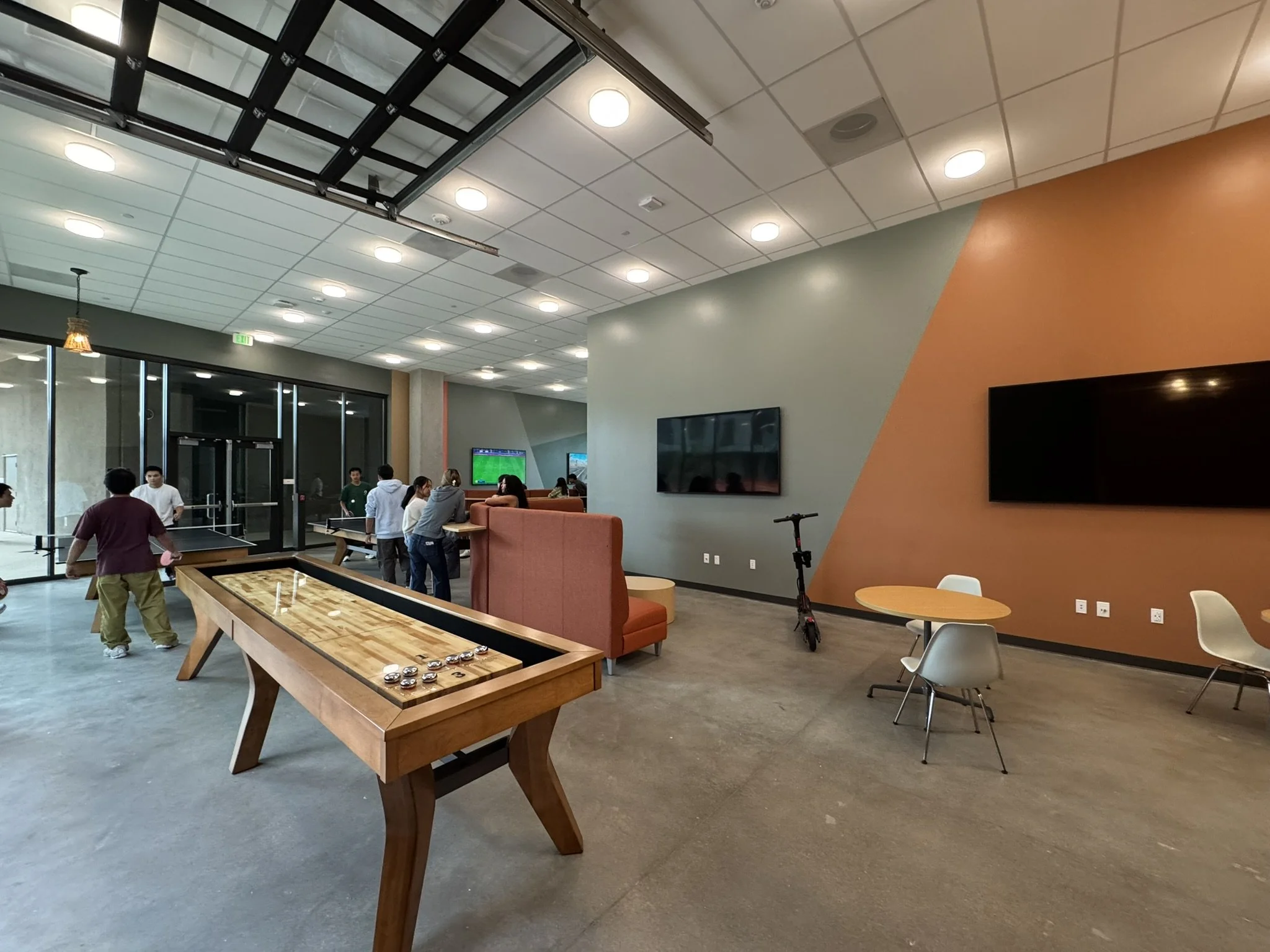 Indoor recreational lounge with tables, chairs, wall-mounted televisions, and a ping pong table. Several people are gathered, with some standing and others sitting.