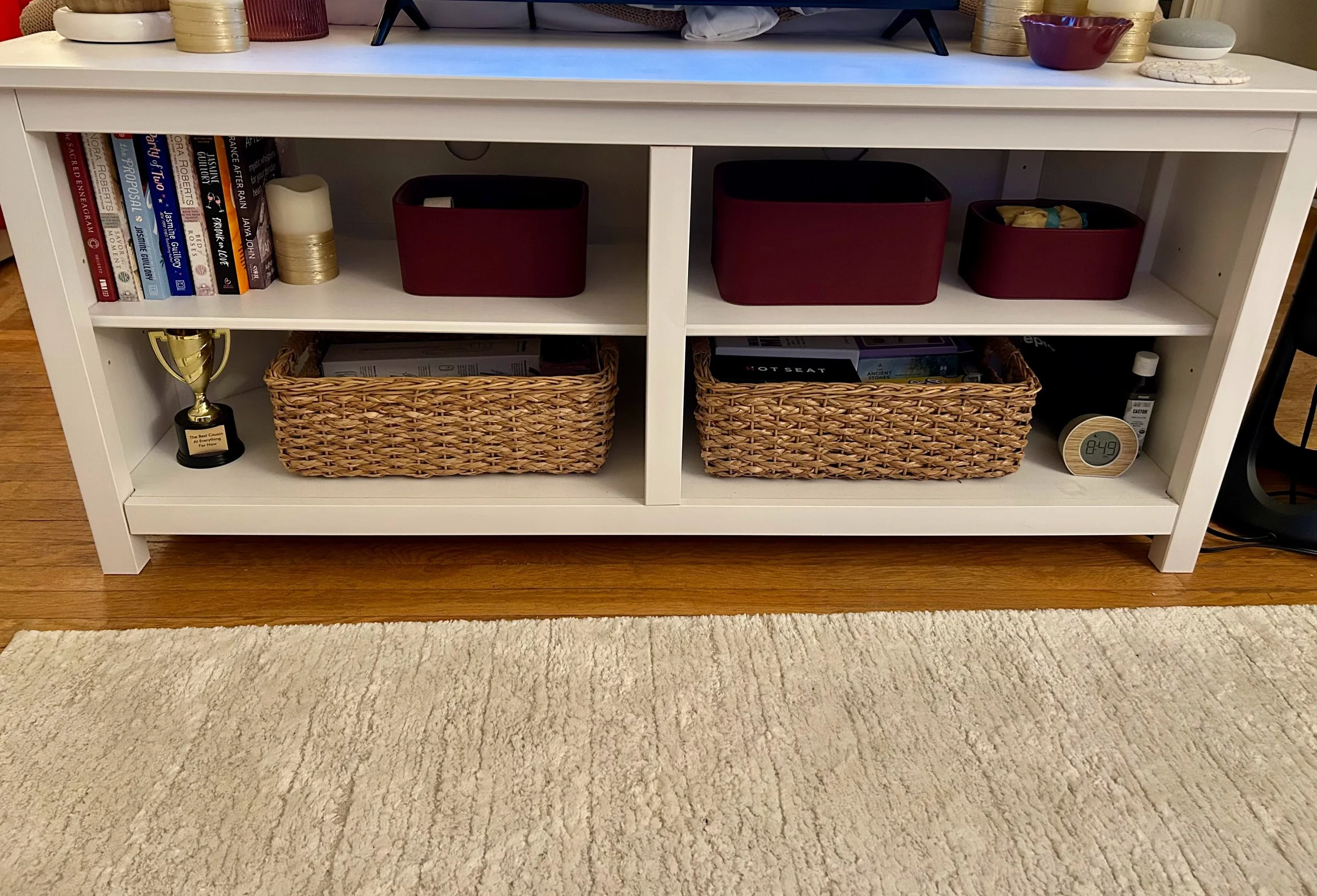 White bookshelf with books, two red storage bins, two woven baskets, a trophy, and various decorative objects and candles on top, against a beige wall on a wooden floor and a beige carpet.