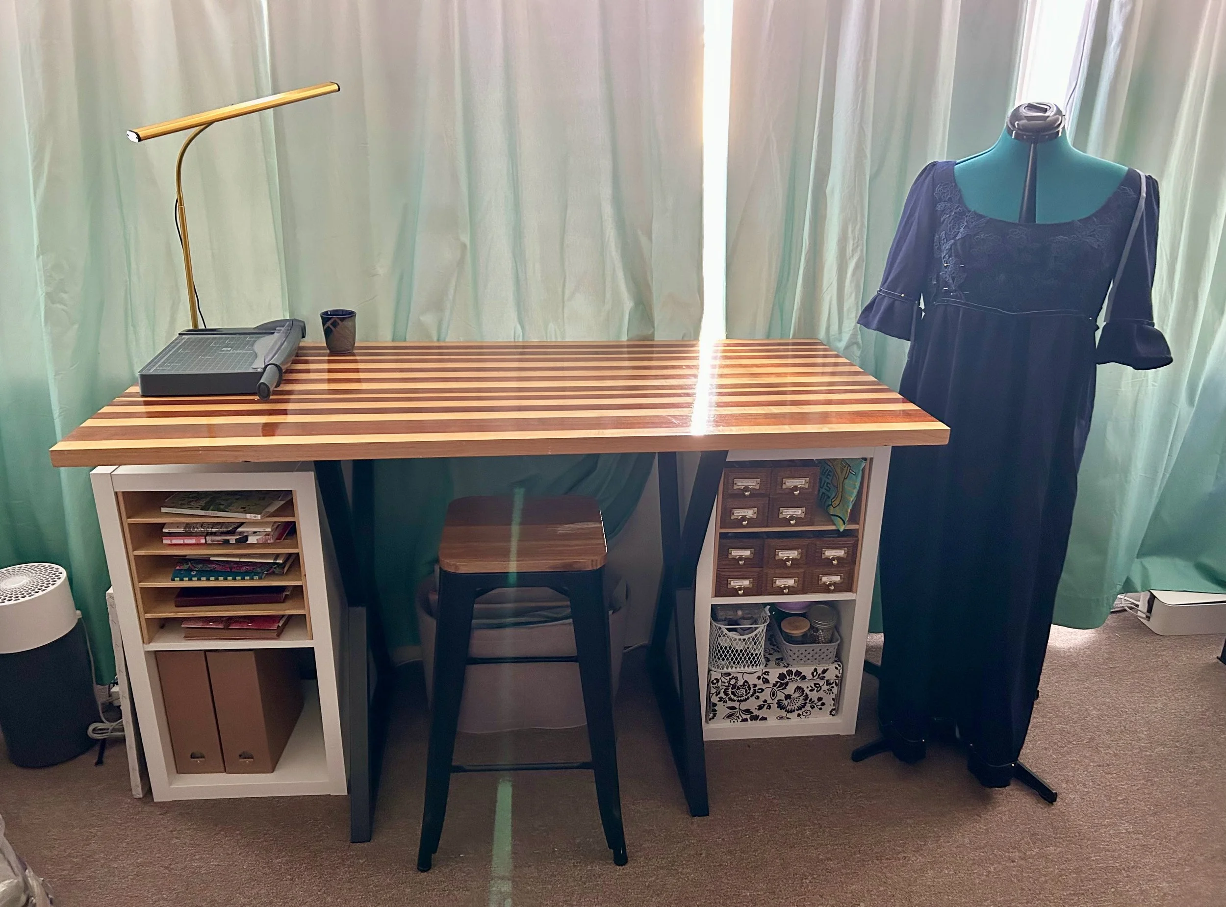 A workspace with a wooden desk, a small black cup, a gold and white desk lamp, and a mannequin wearing a navy blue dress. There is a bookshelf underneath the desk and a green curtain in the background.