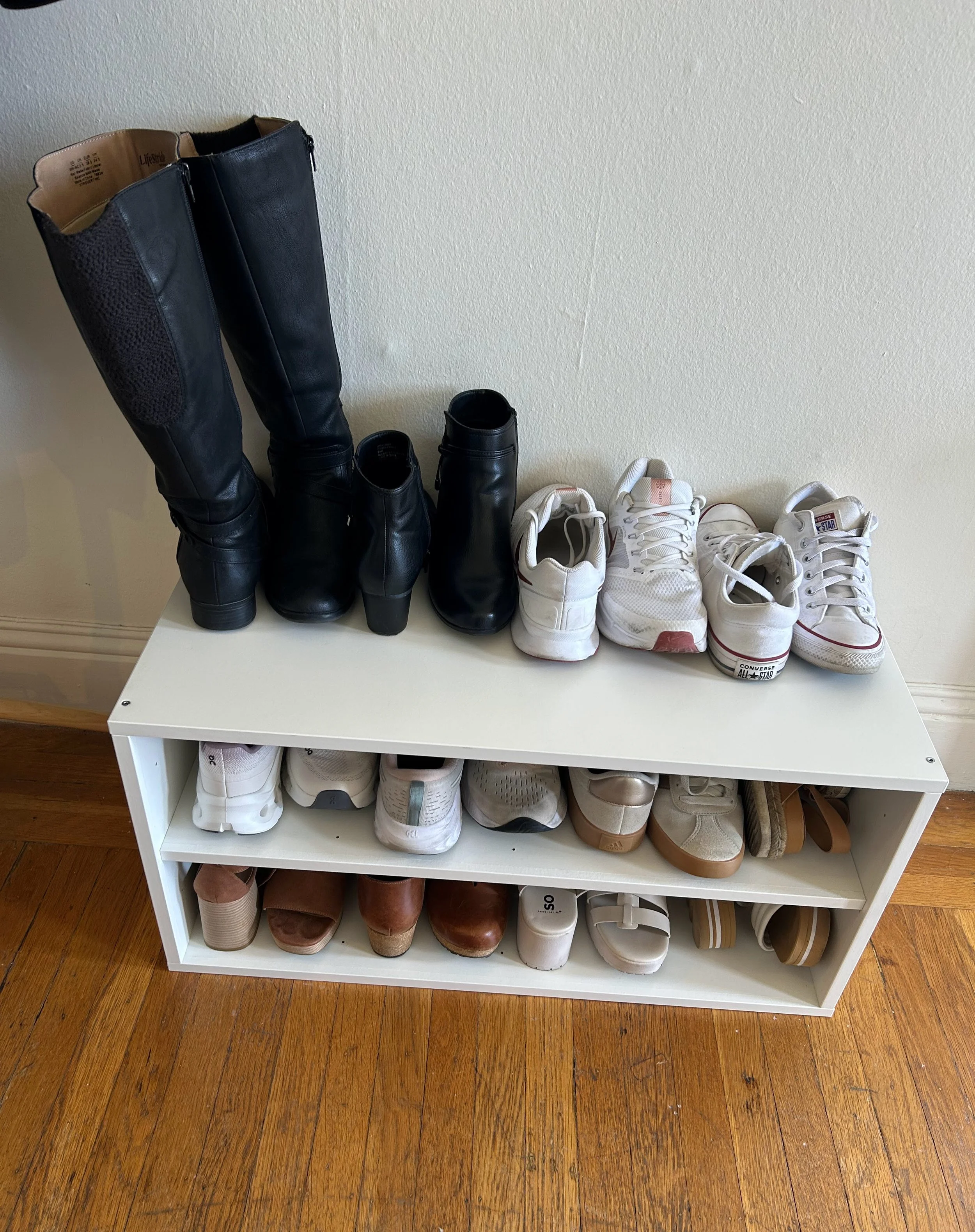 A white shoe rack with two shelves filled with various shoes, positioned against a wall with a wooden floor beneath it.