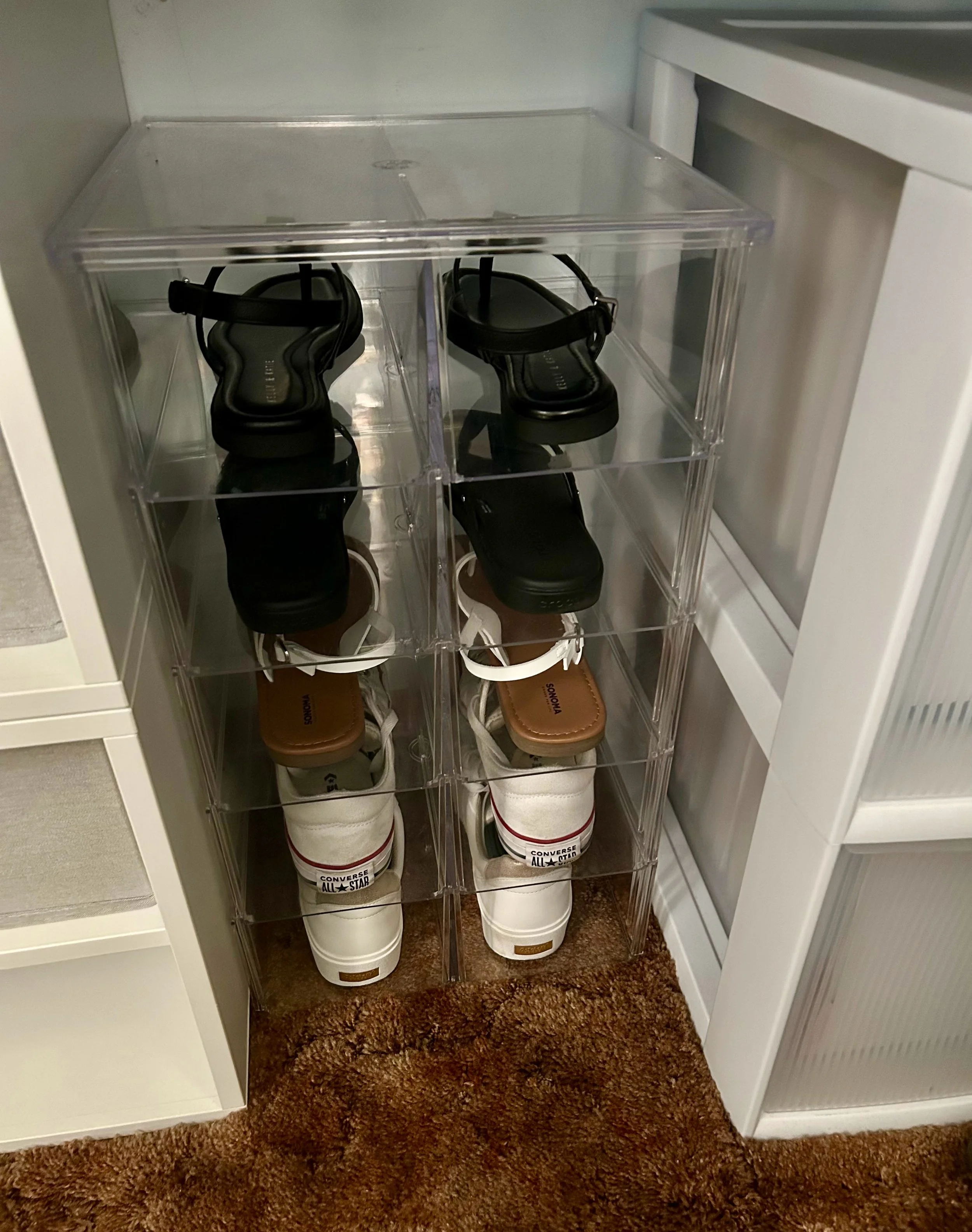 A set of transparent plastic shoe storage drawers containing black sandals, brown sneakers, and white high-top sneakers. The drawers are placed inside a white cabinet on a brown carpeted floor.