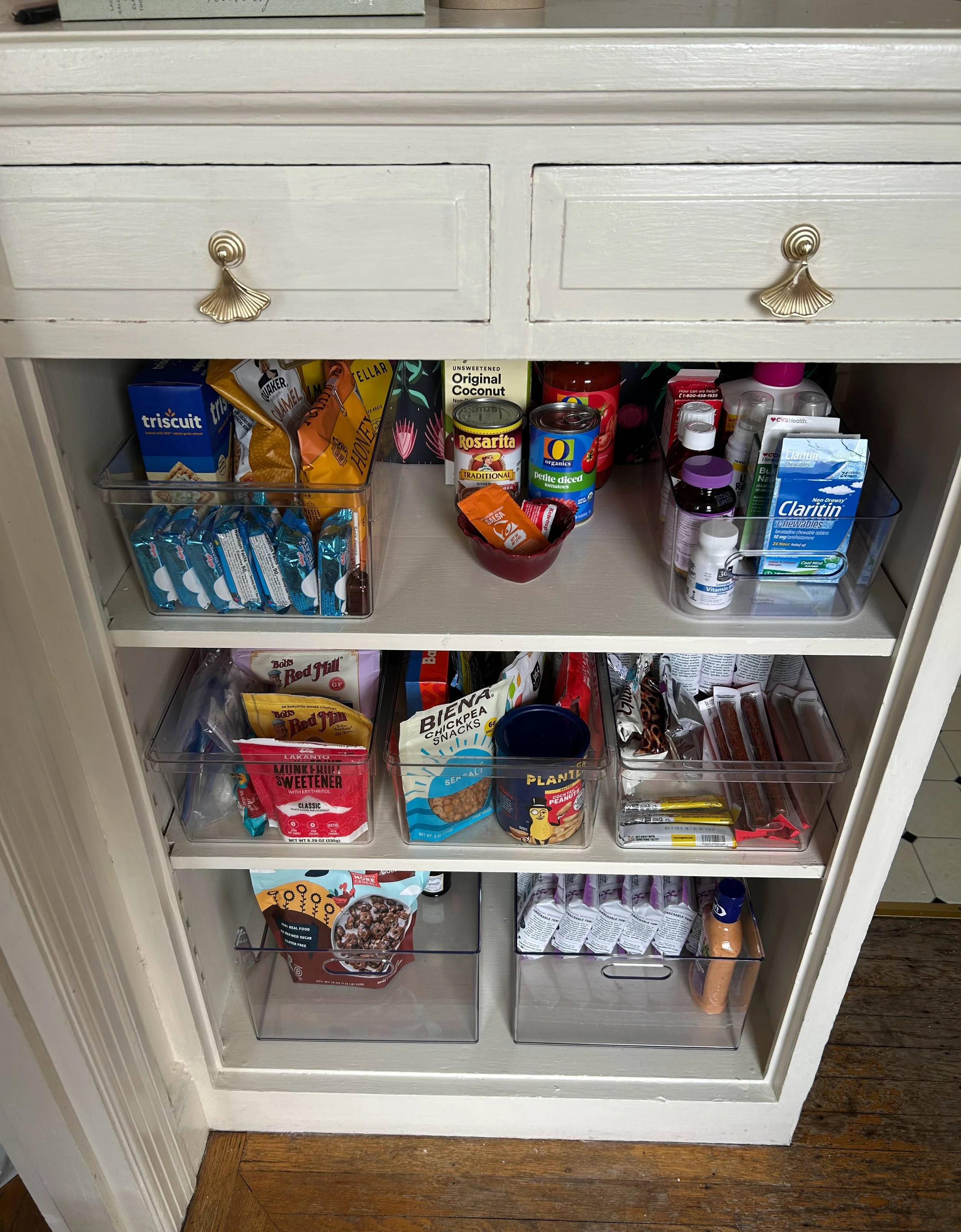 A white kitchen cabinet with three shelves. The middle shelf has two clear plastic containers filled with snack items including granola bars, trail mix, and cracker packets. The open shelves contain various canned goods, boxes of snacks, and personal