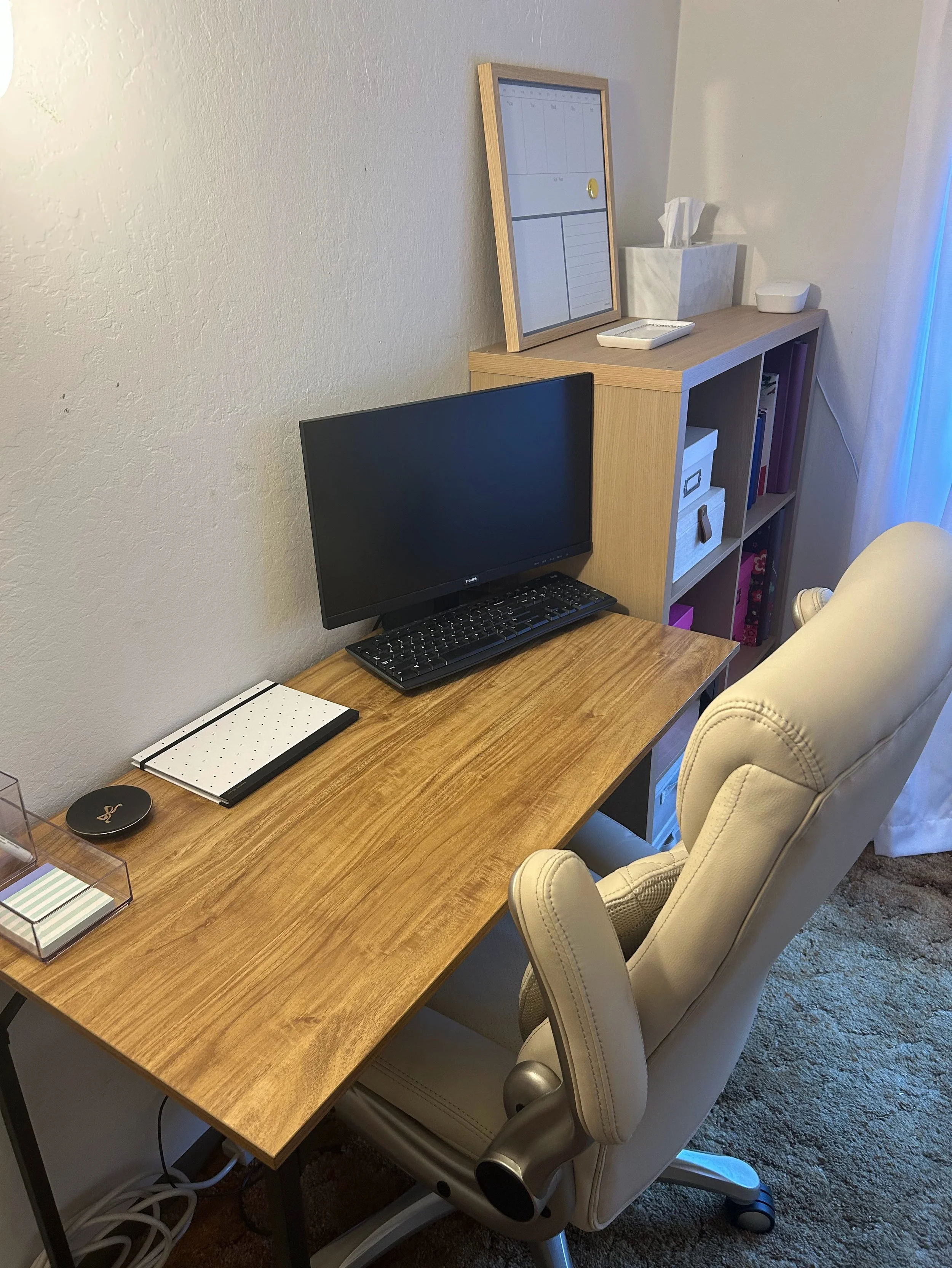 A home office setup with a wooden desk, a computer monitor, a keyboard, and a notebook. Behind the desk is a beige office chair. On the wall, there is a bookshelf with binders and a tissue box. On top of the bookshelf, there is a framed whiteboard or