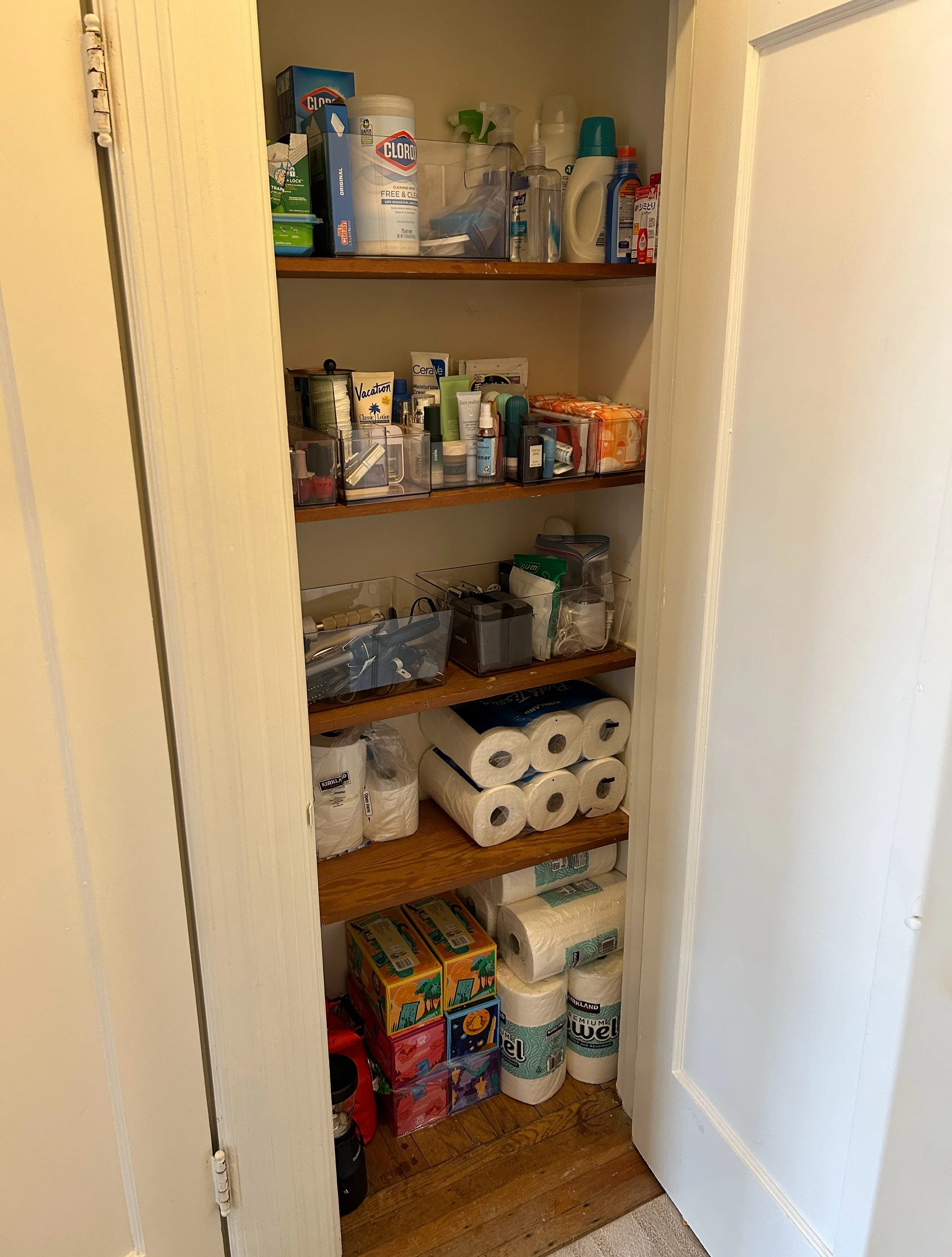 A pantry closet with multiple shelves containing household supplies including toilet paper, paper towels, cleaning products, toiletries, and boxed items.