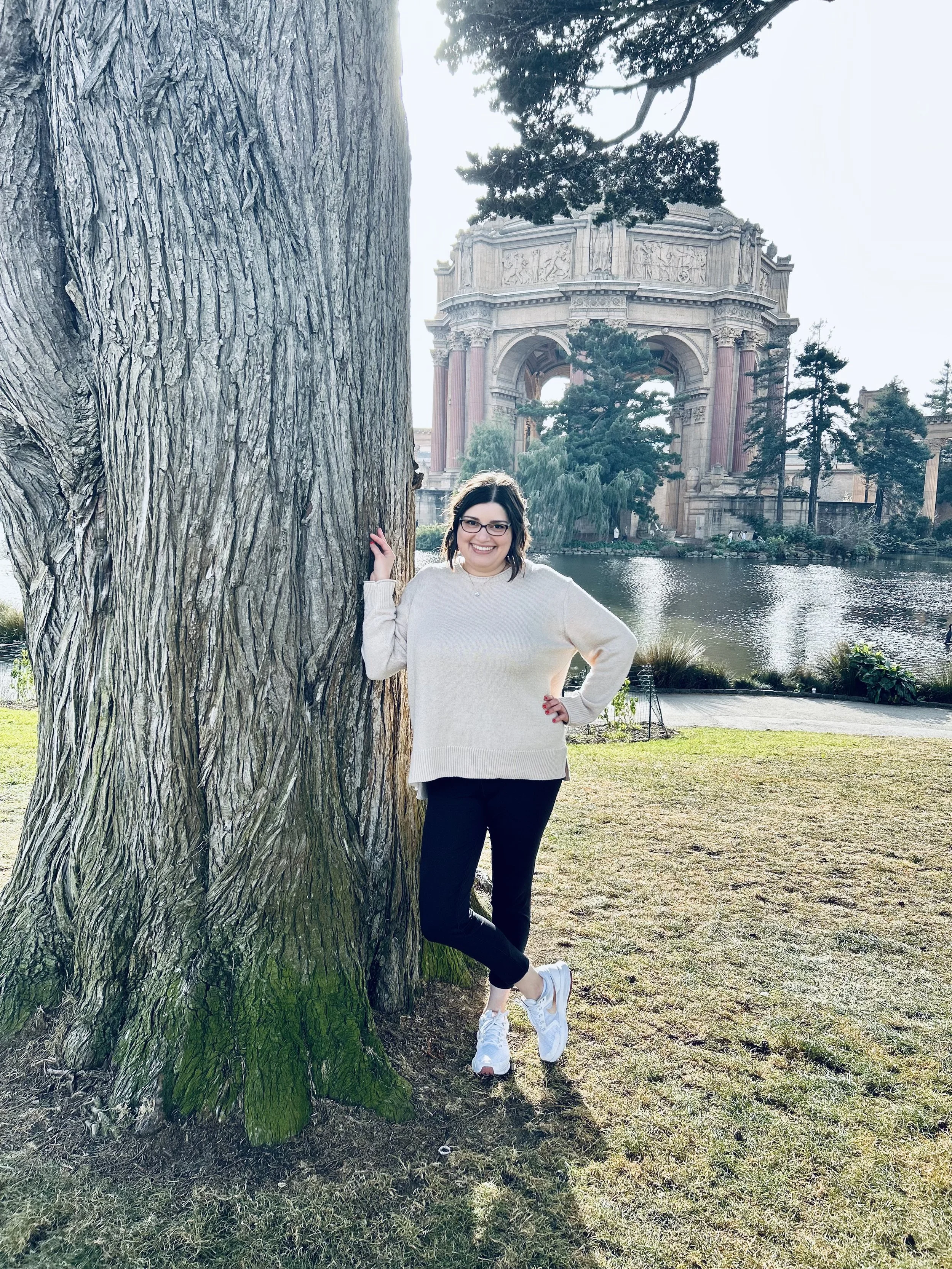 A woman with glasses smiling and posing outdoors next to a large tree with a historic arch monument in the background