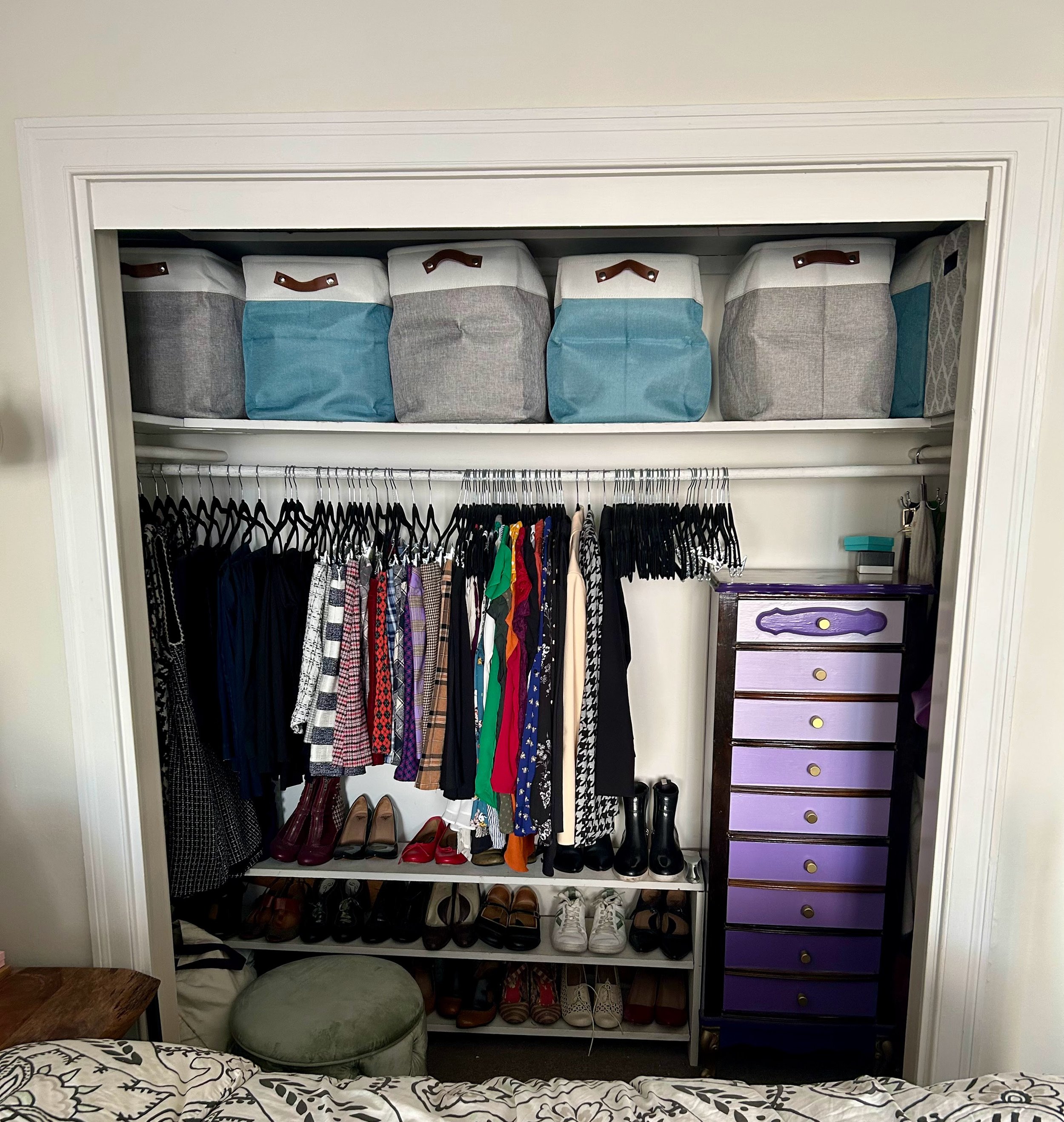 Open closet with shoes on bottom shelf, hanging clothes on middle rod, storage bins on top shelf, purple dresser to the right, and various accessories at the bottom right corner.