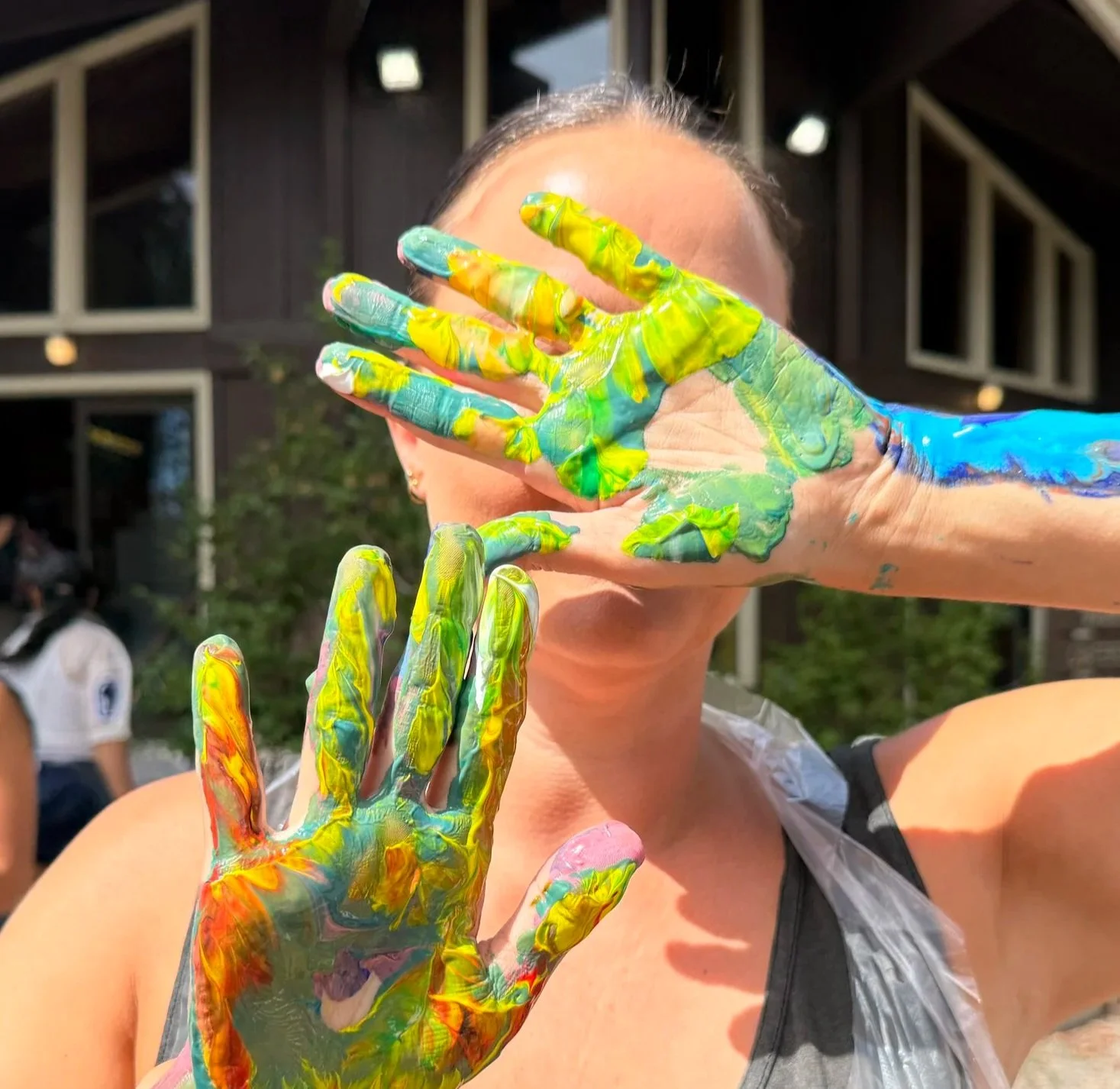 A person with colorful paint on their hands and face covering part of their face, outdoors near a building with triangular windows.