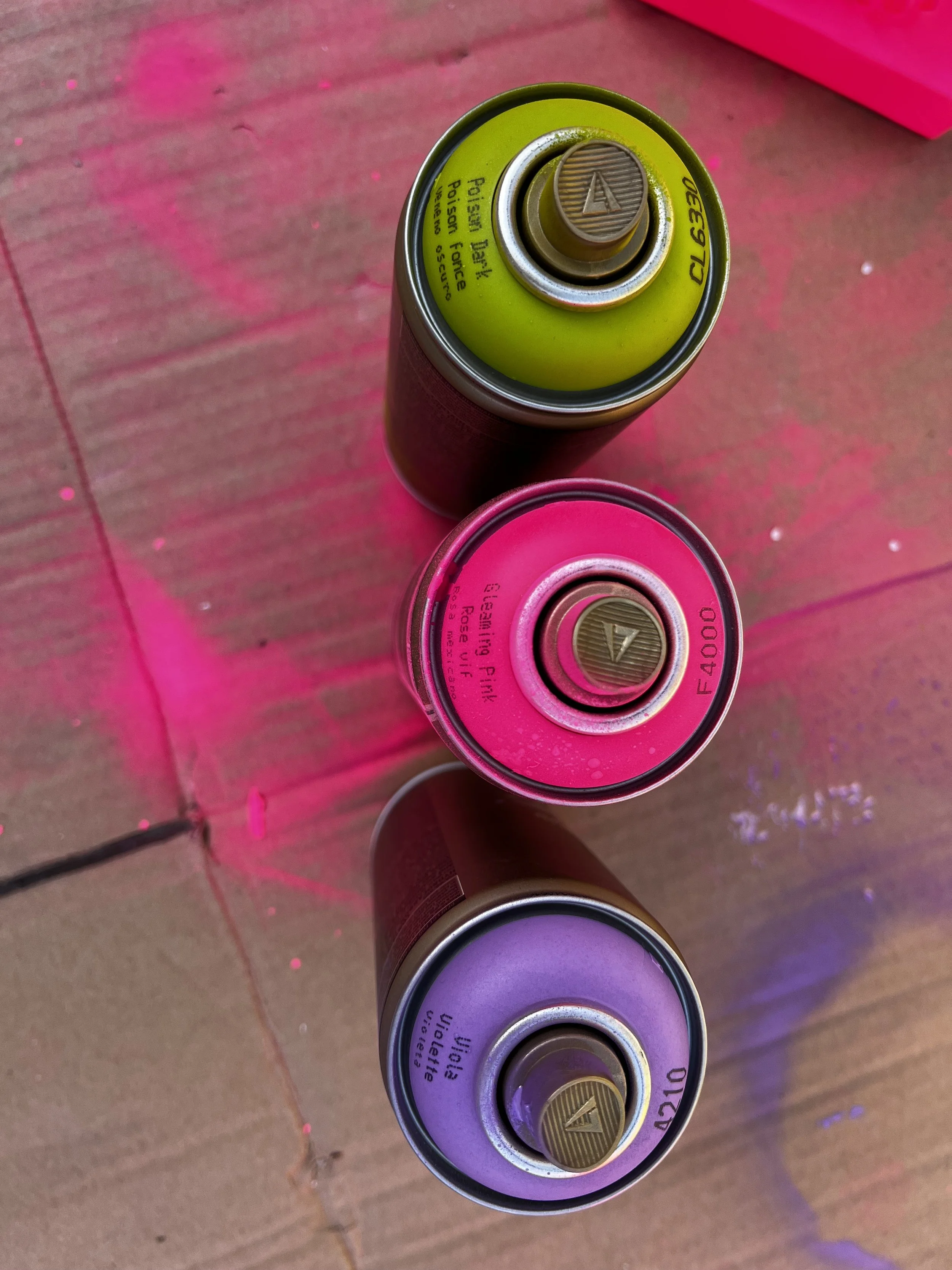 Three spray paint cans in different colors: green at the top, pink in the middle, and purple at the bottom, positioned on a wooden surface under pink and purple lighting.