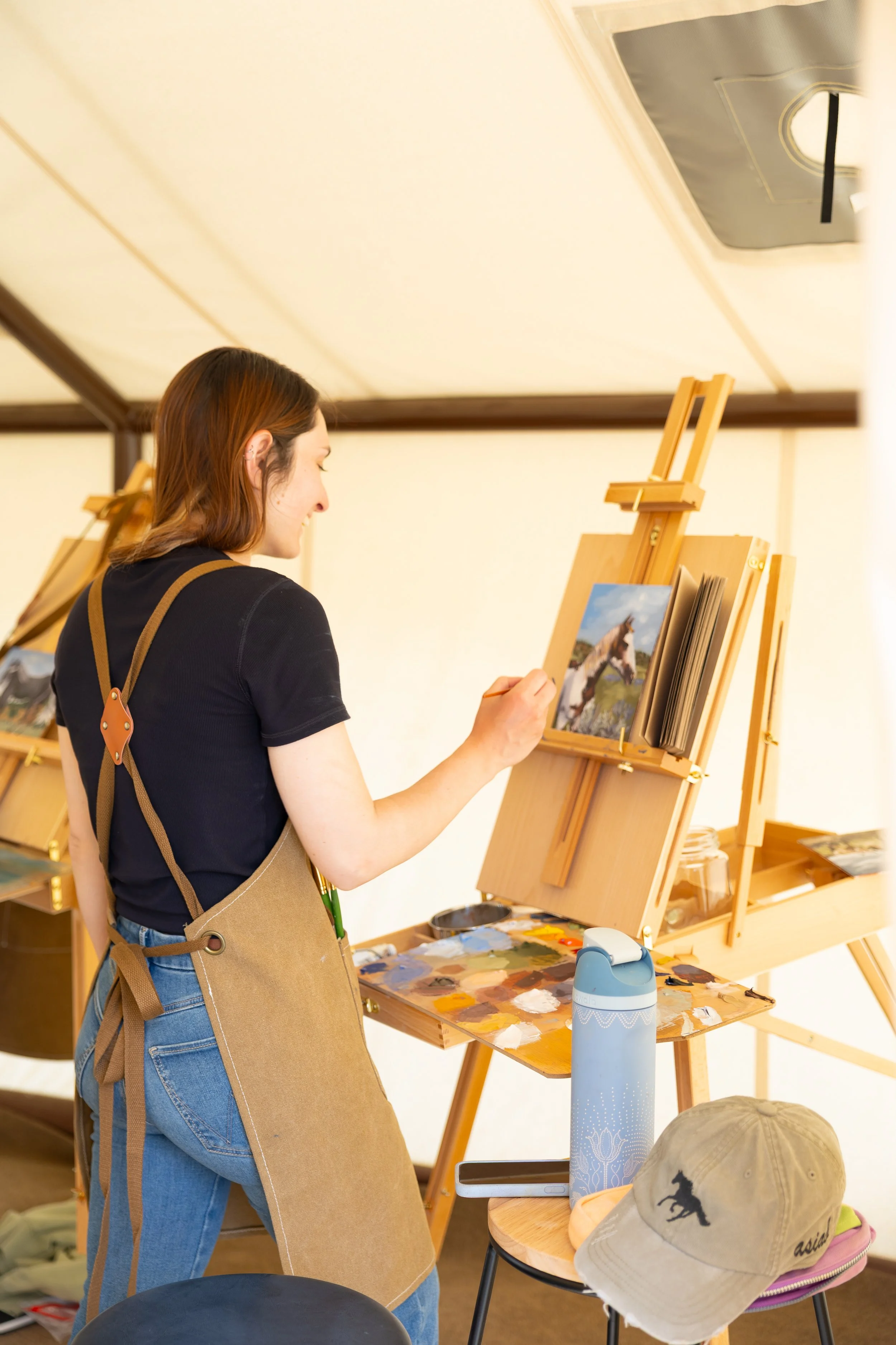 A woman painting a horse on canvas at an art easel, wearing a beige apron and looking at her artwork, with art supplies and accessories on the table and a beige baseball cap with a horse logo in the foreground.