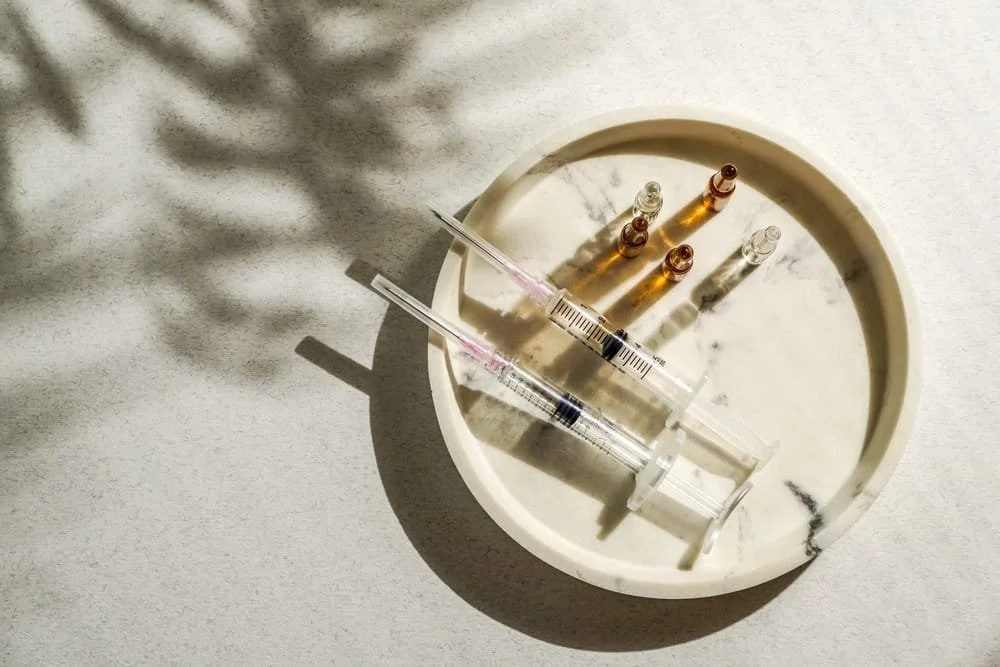A round white dish with five small glass vials and a syringe, with shadows cast on a light surface.